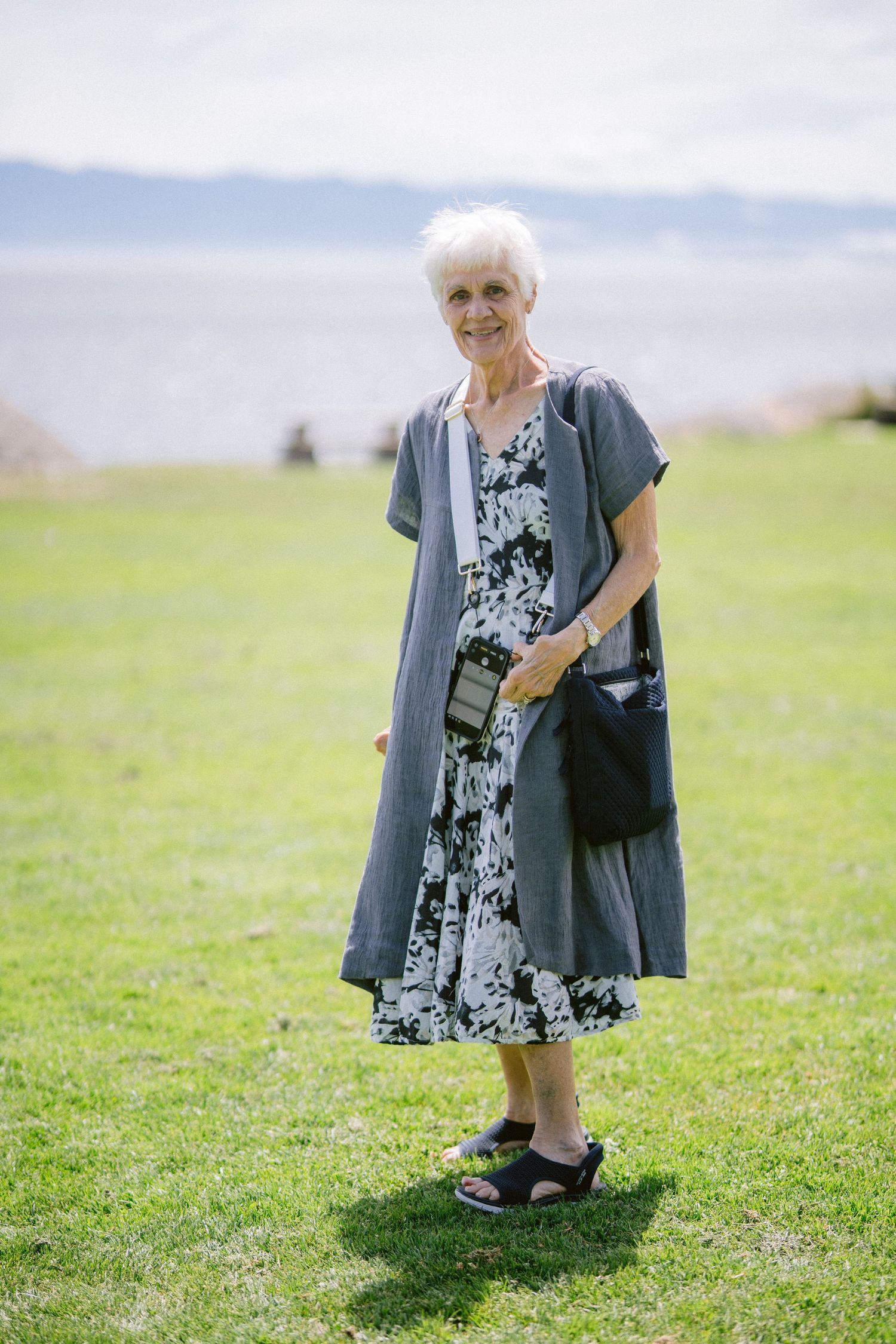 Elderly woman in floral dress and gray cardigan smiling outdoors on green grass near waterfront.