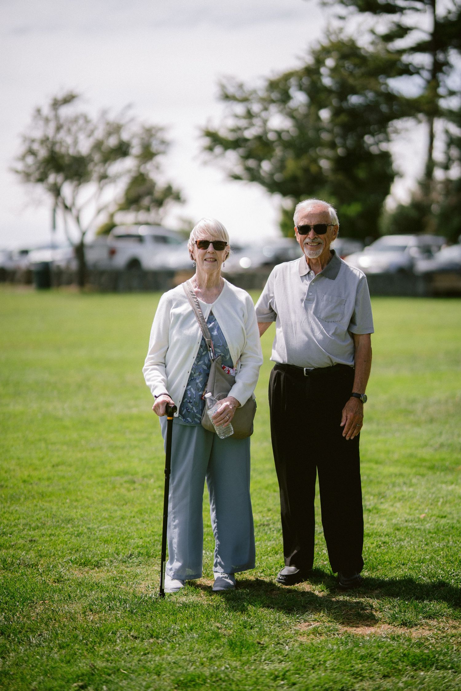 Senior couple smiling outdoors on grass, woman with cane in white cardigan and man in gray polo shirt.