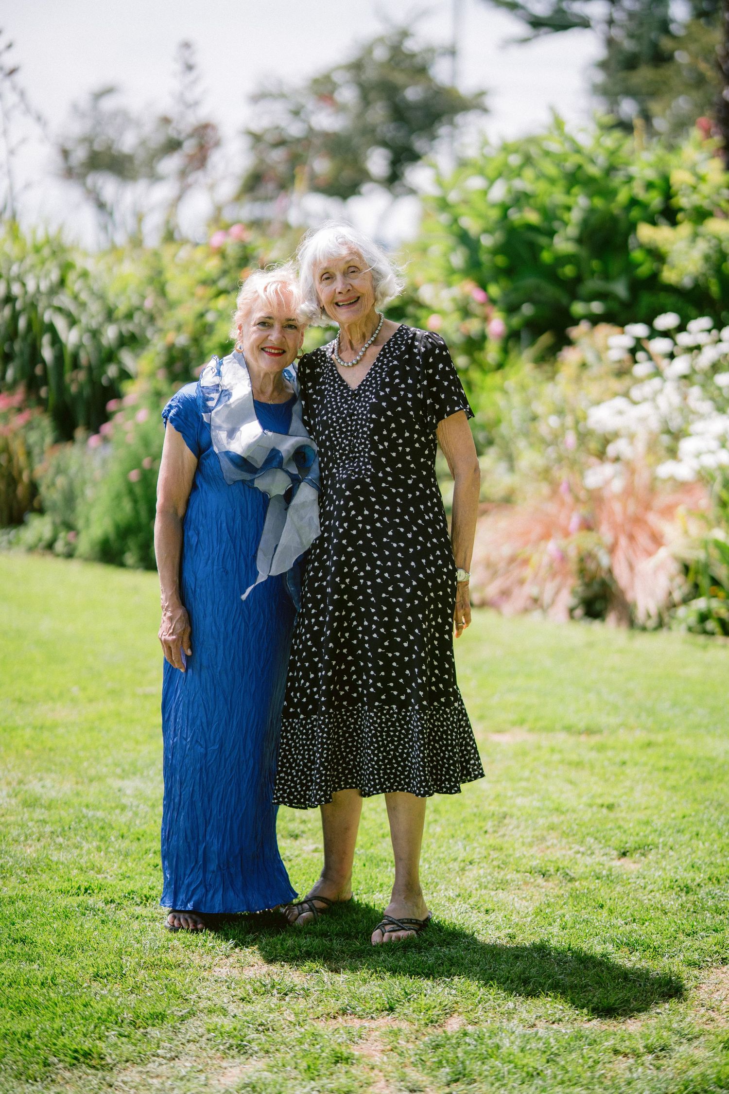 Two elegant senior women posing together in a lush garden, one in blue dress, one in black polka dot dress.