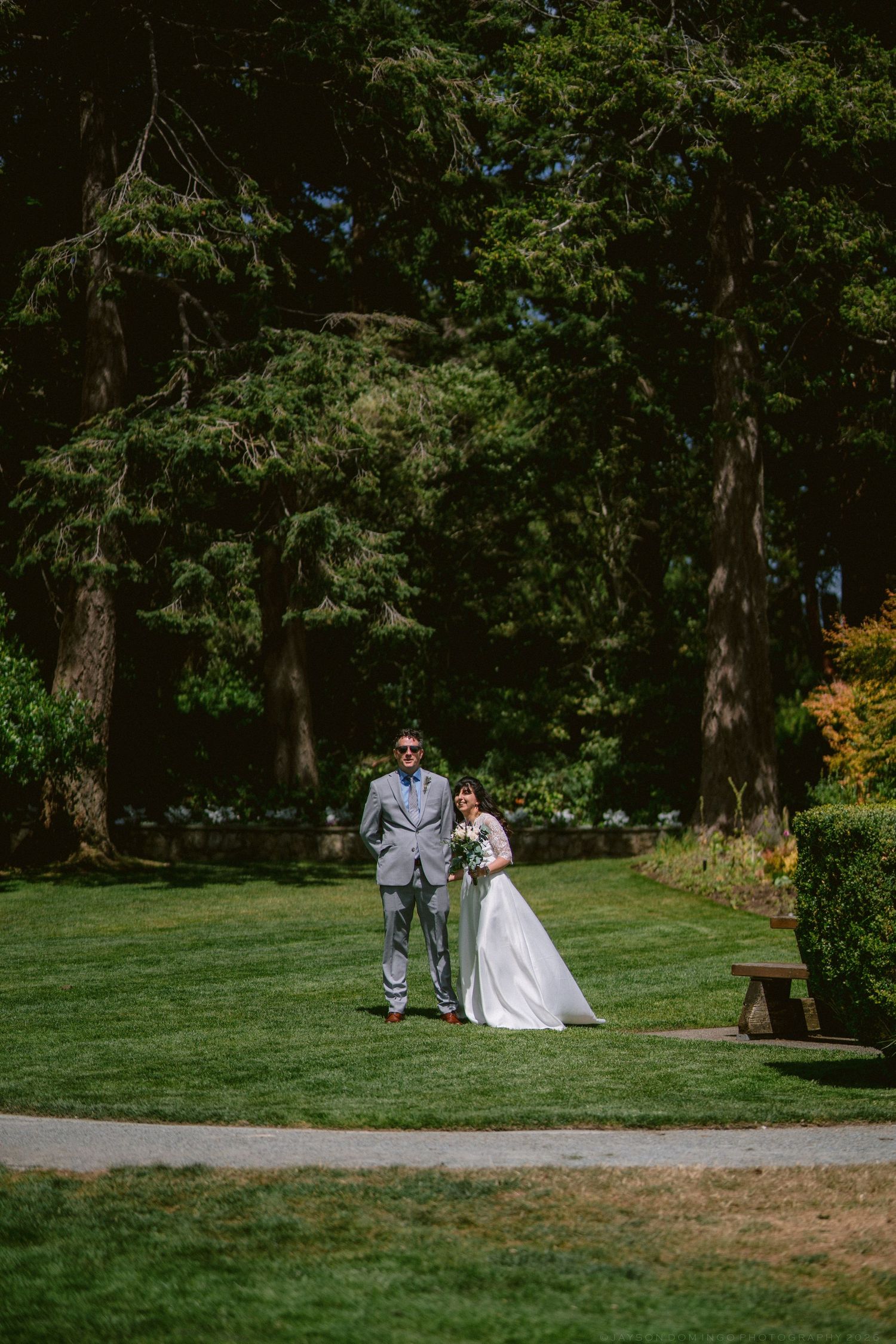 Bride and groom walking together on a sunlit green lawn surrounded by tall trees.
