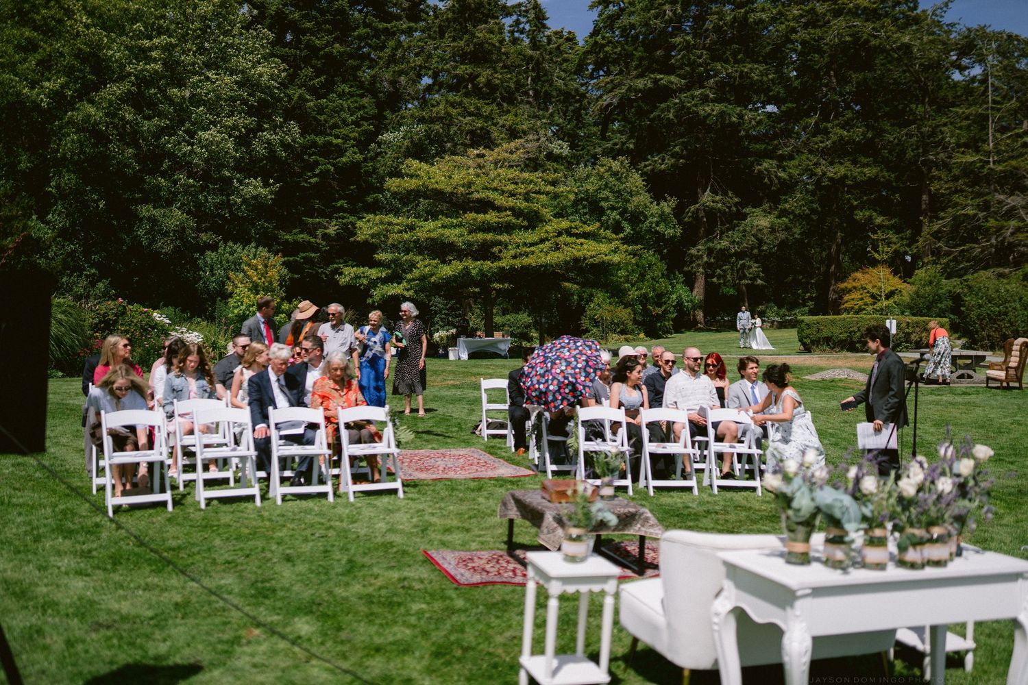 Outdoor wedding ceremony on green lawn with white chairs, guests seated, surrounded by lush trees on a sunny day.