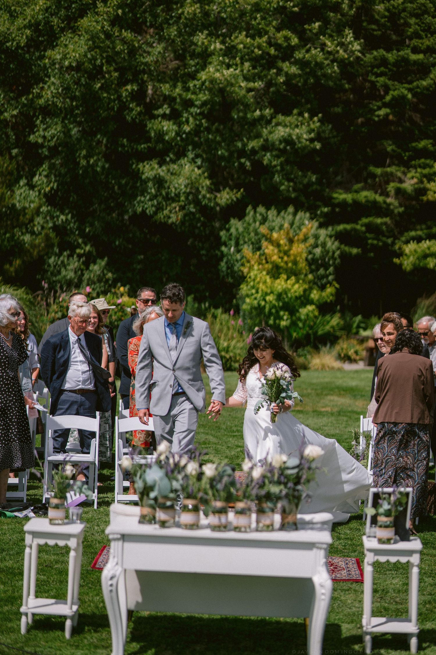 Bride and groom walking down the aisle at an outdoor garden wedding ceremony with guests seated.