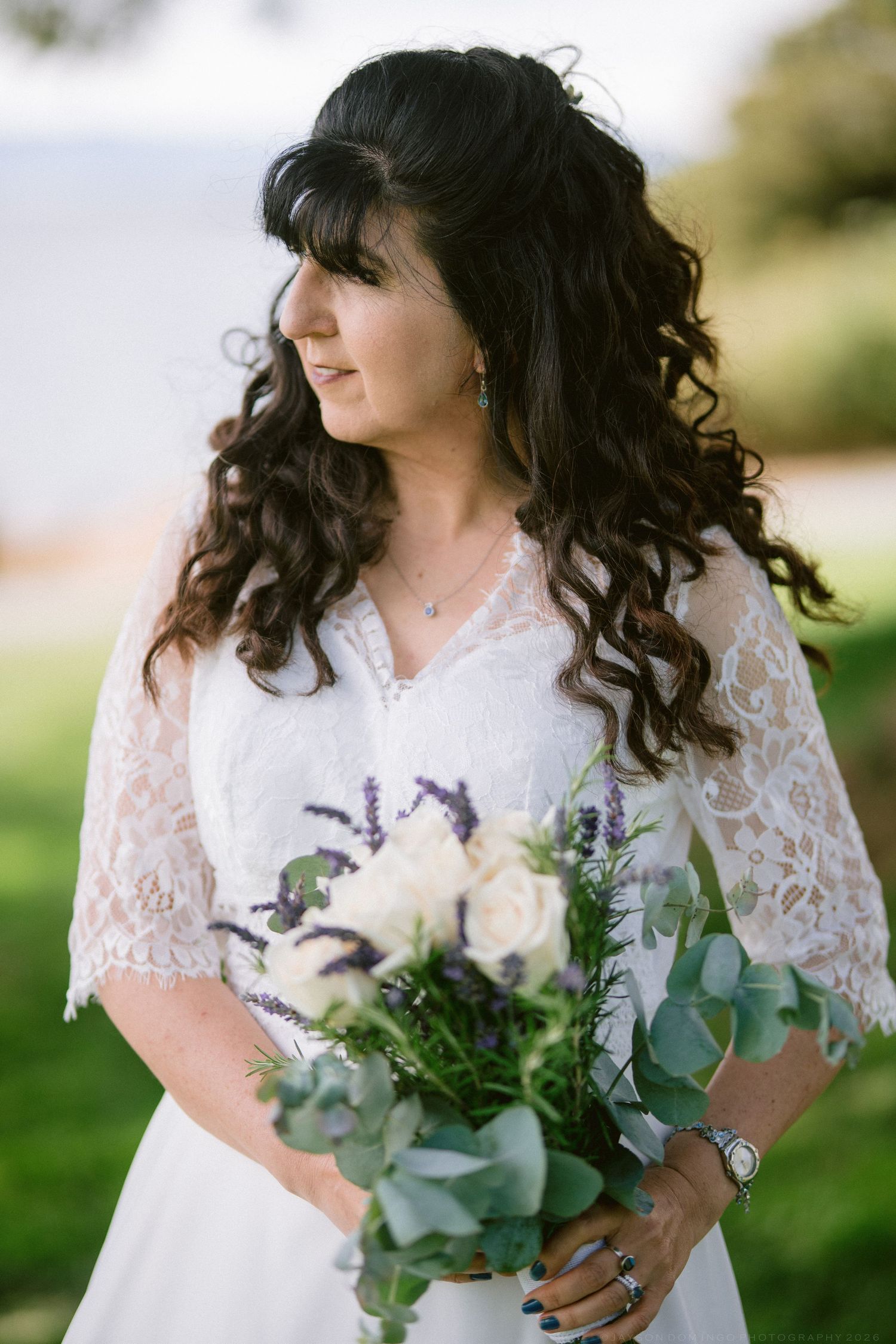 Bride with curly dark hair holding white and lavender bouquet in lace wedding dress outdoors.