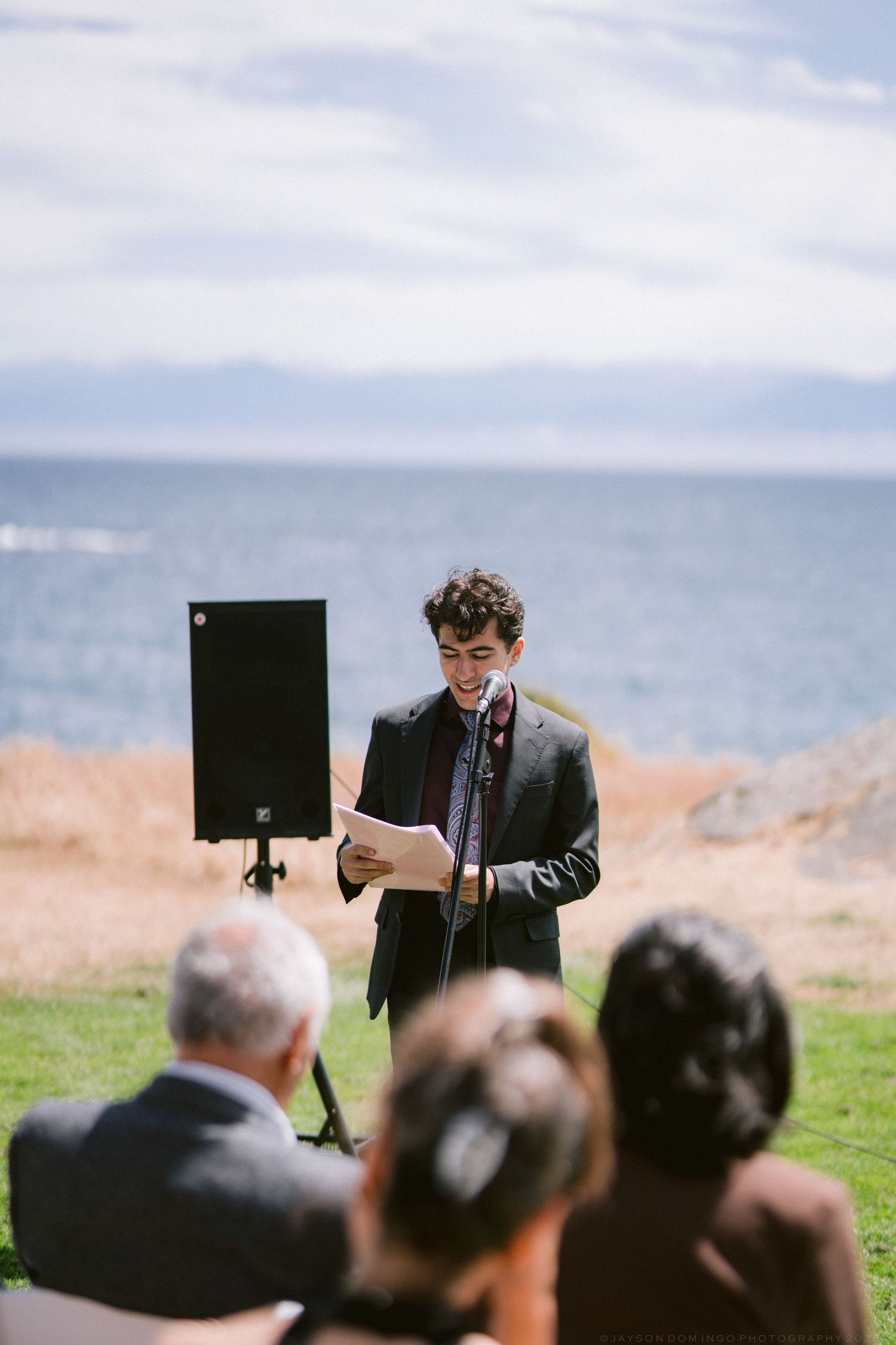 Young man in dark suit reading vows at outdoor wedding ceremony with ocean backdrop and seated guests.