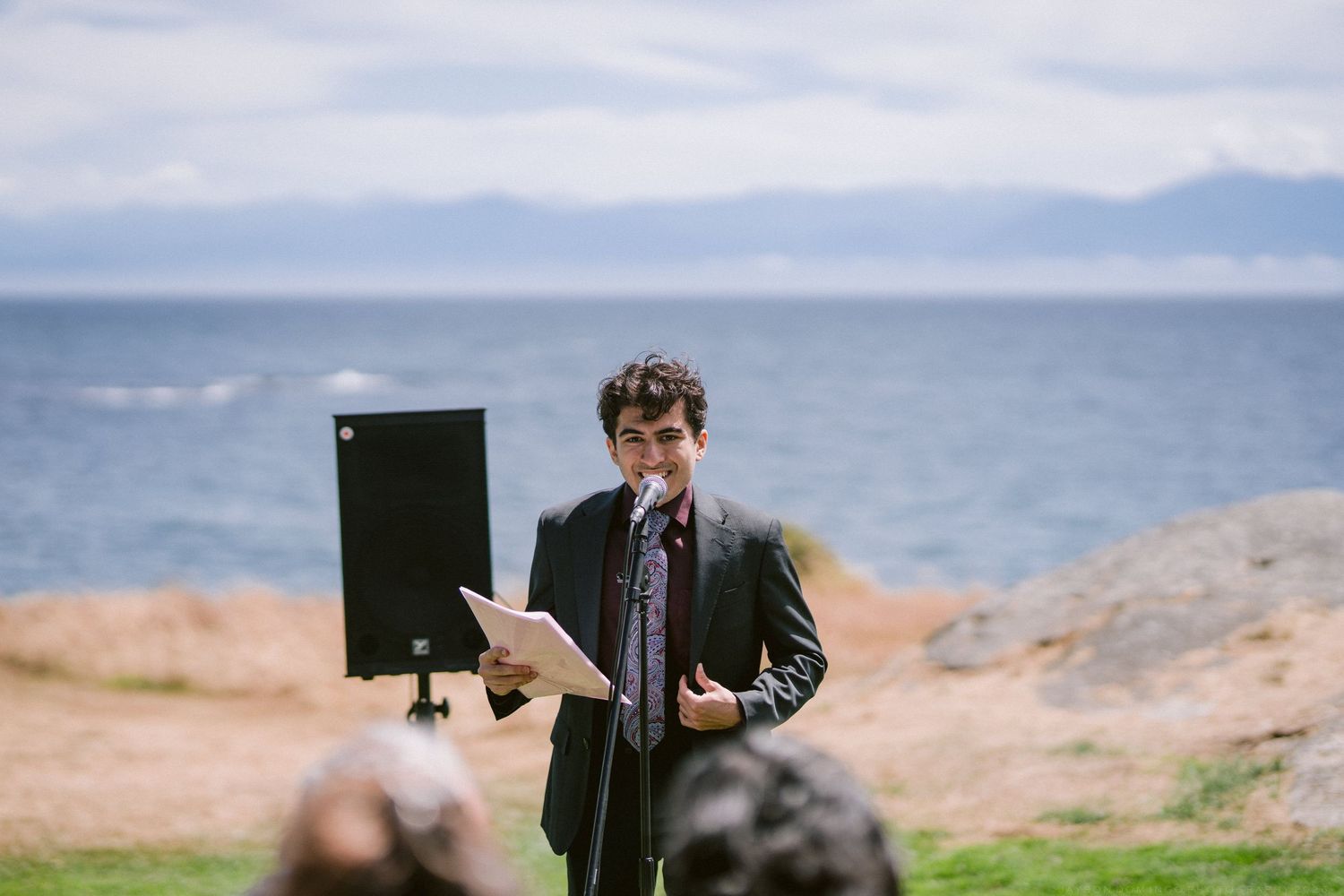 Man in suit speaking at outdoor waterfront ceremony with microphone stand and scenic ocean backdrop.