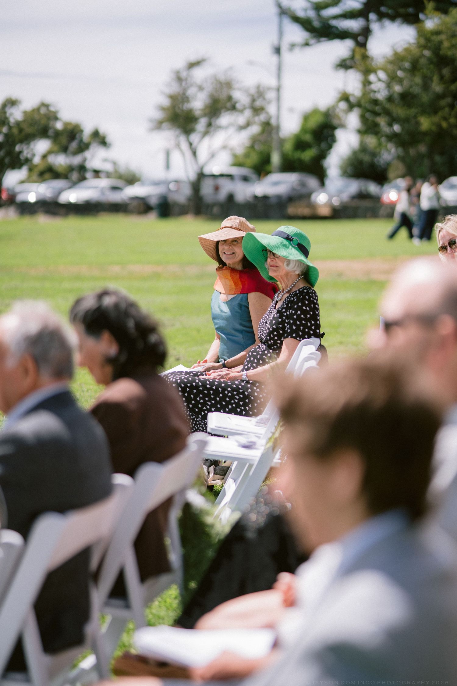 Guests seated outdoors at a sunny wedding ceremony, two women wearing colorful wide-brim hats in the foreground.