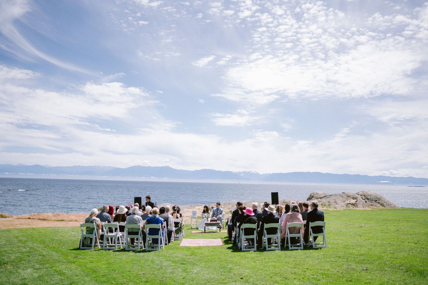 Outdoor wedding ceremony on green lawn overlooking a serene lake with mountains and blue sky backdrop.