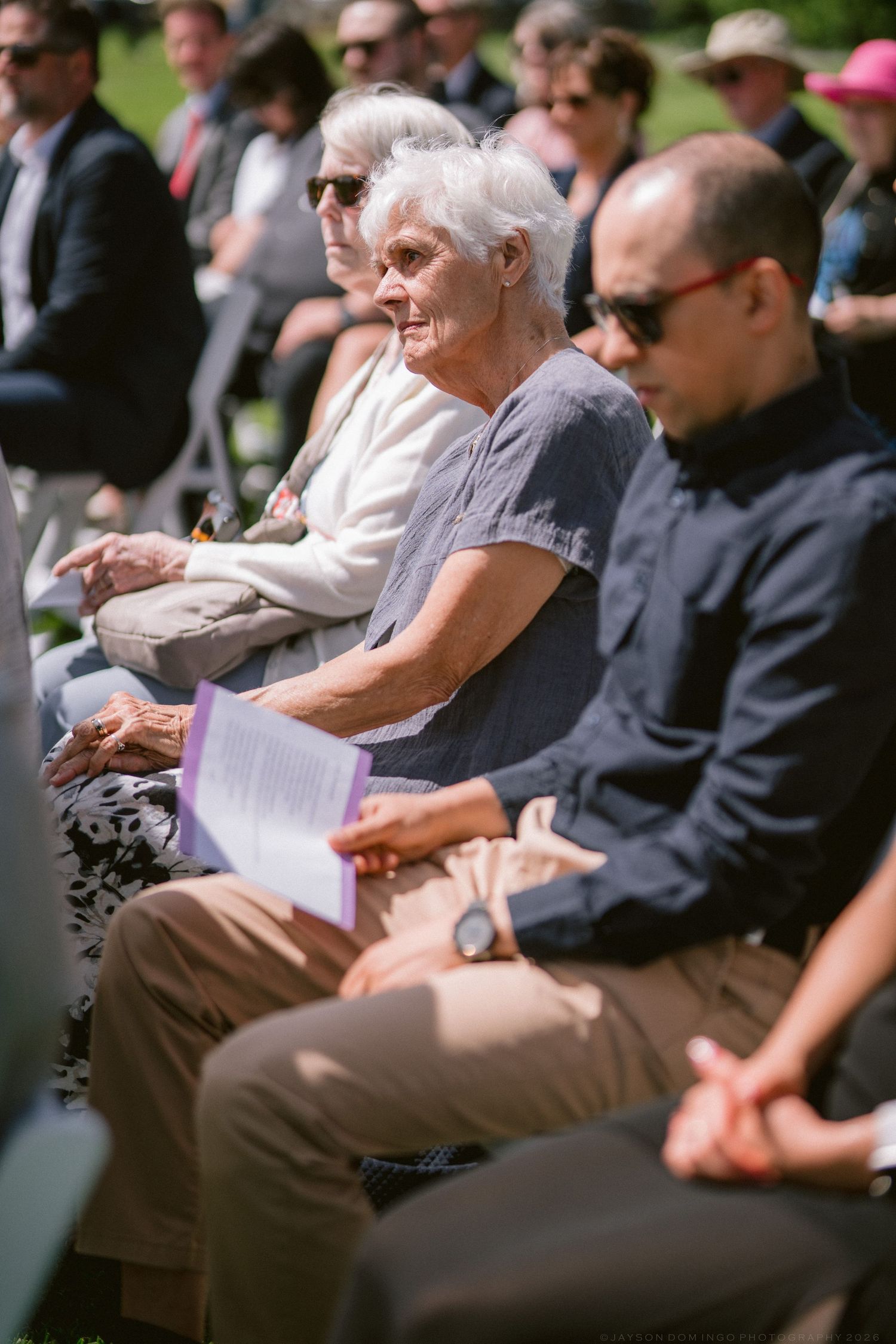 Elderly guests seated outdoors at a sunny wedding ceremony, woman in gray holding purple program.
