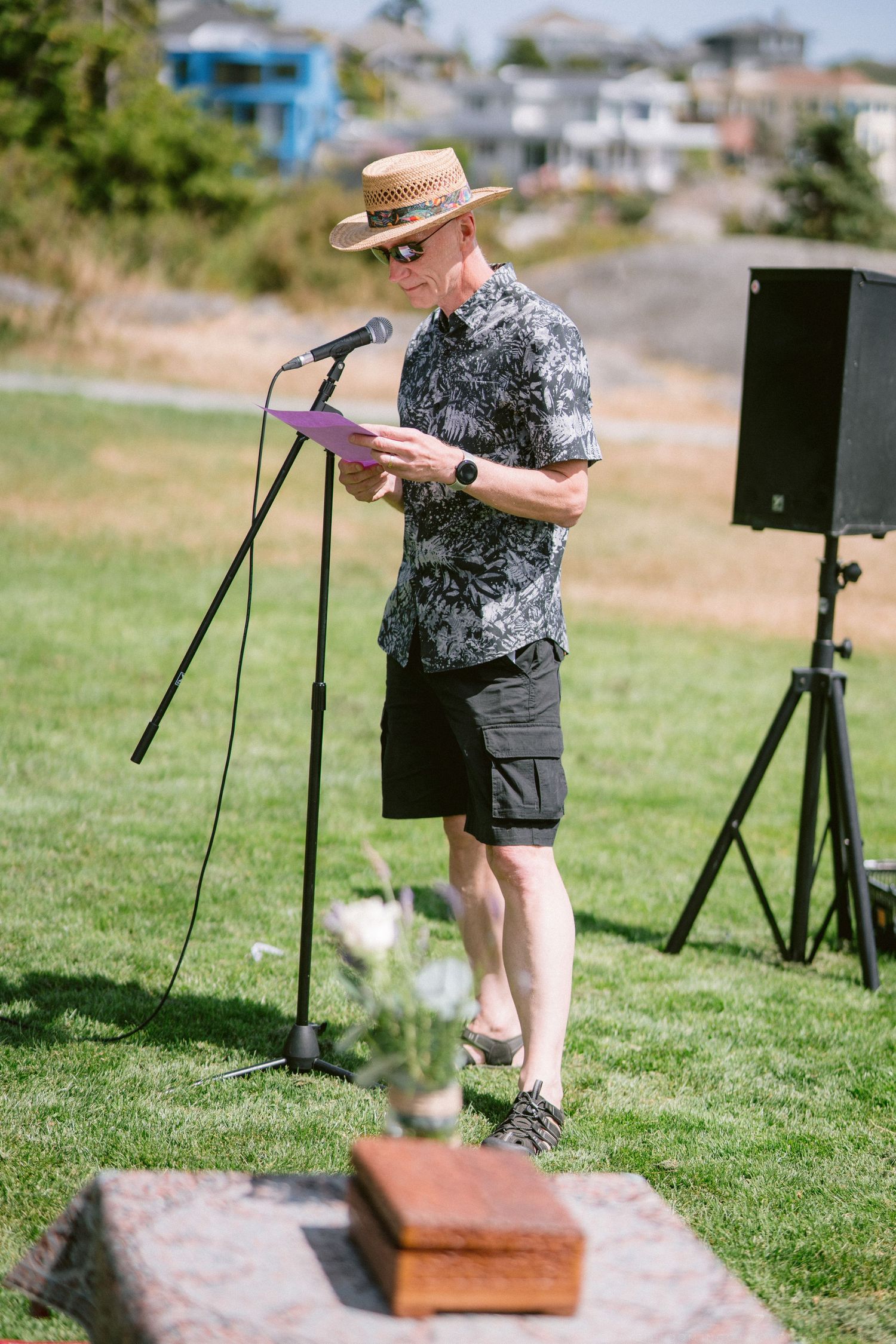 Man in straw hat and floral shirt reading at microphone stand during outdoor wedding ceremony.