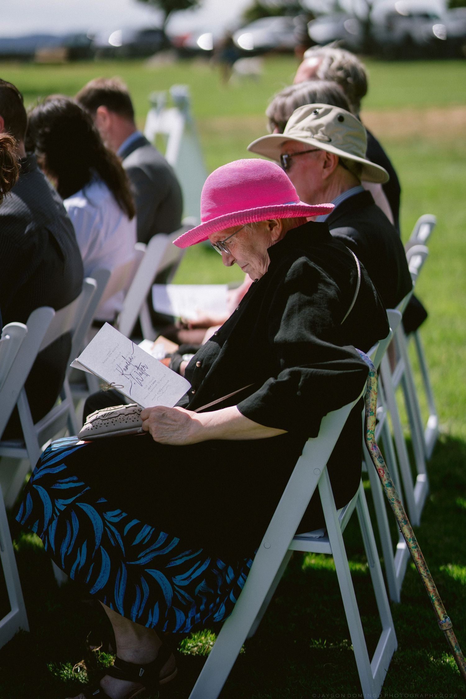 Senior woman in pink sun hat reading program at outdoor wedding ceremony seated in white chairs.