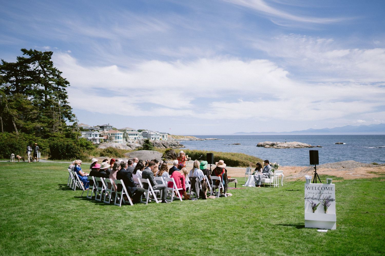 Outdoor wedding ceremony on green lawn overlooking rocky coastline with guests seated in white chairs under blue sky.