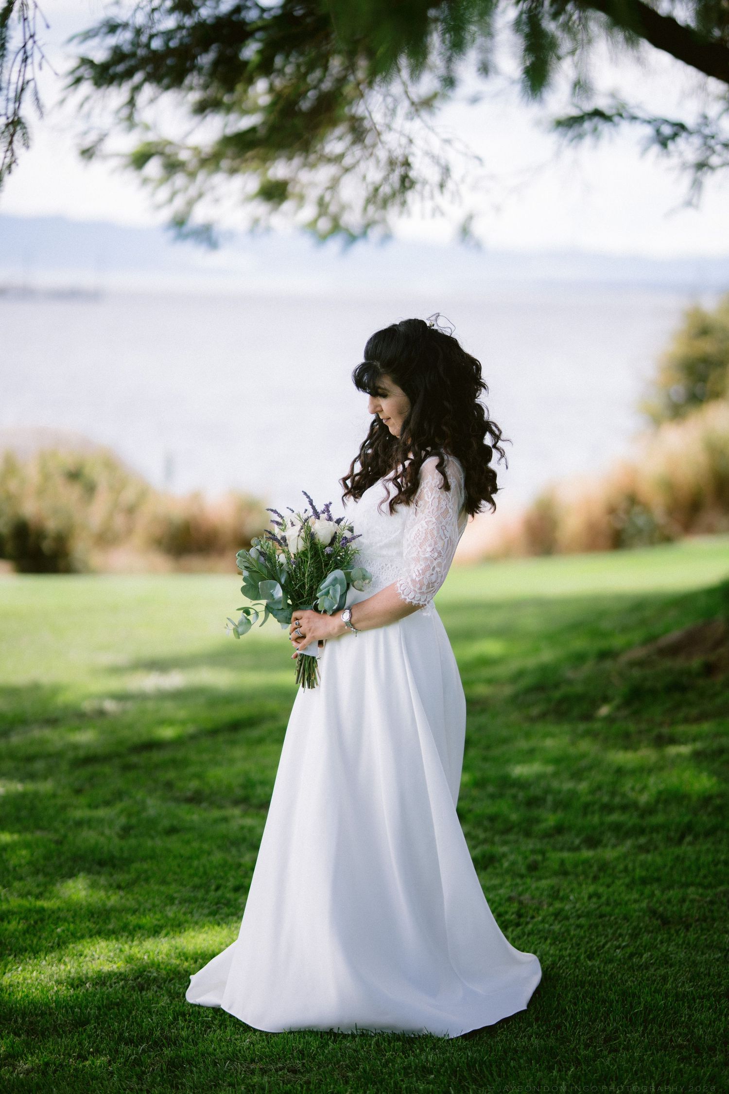 Bride in white lace gown holding a green bouquet stands on grass near a waterfront.