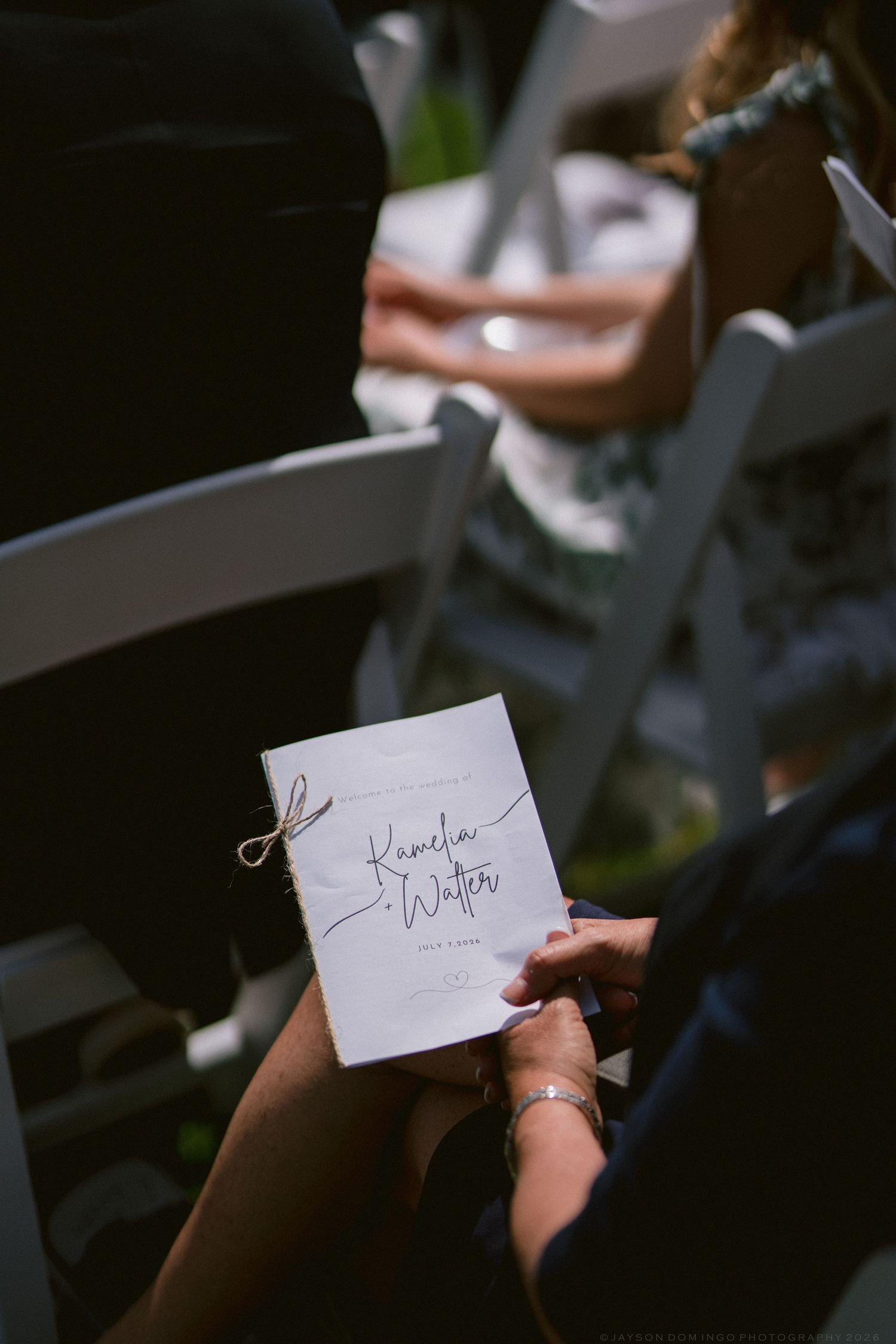 Guest holds elegant wedding program booklet tied with twine at outdoor ceremony.