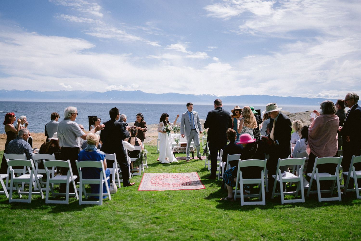 Wedding ceremony on coastal lawn with guests in white chairs as couple exchanges vows with ocean backdrop.