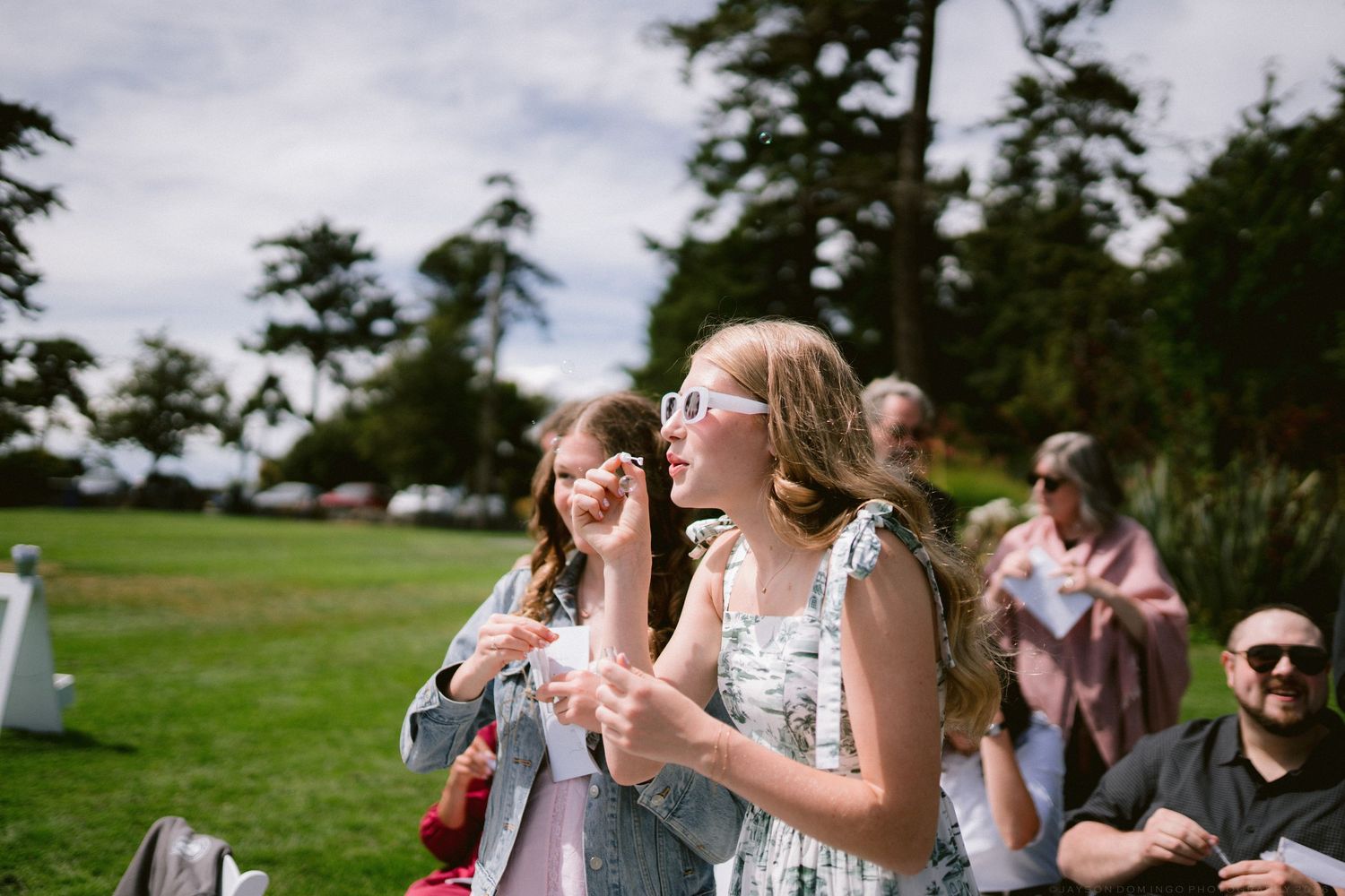 Young girls blowing bubbles at an outdoor wedding reception on a green lawn surrounded by tall trees.