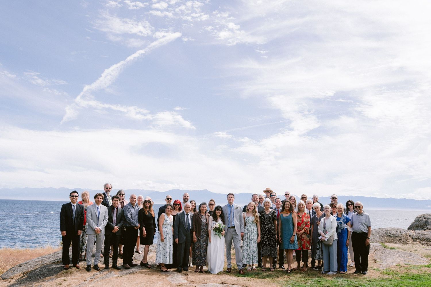 Large wedding group photo outdoors by the ocean on a sunny day with blue skies and rocky shoreline.