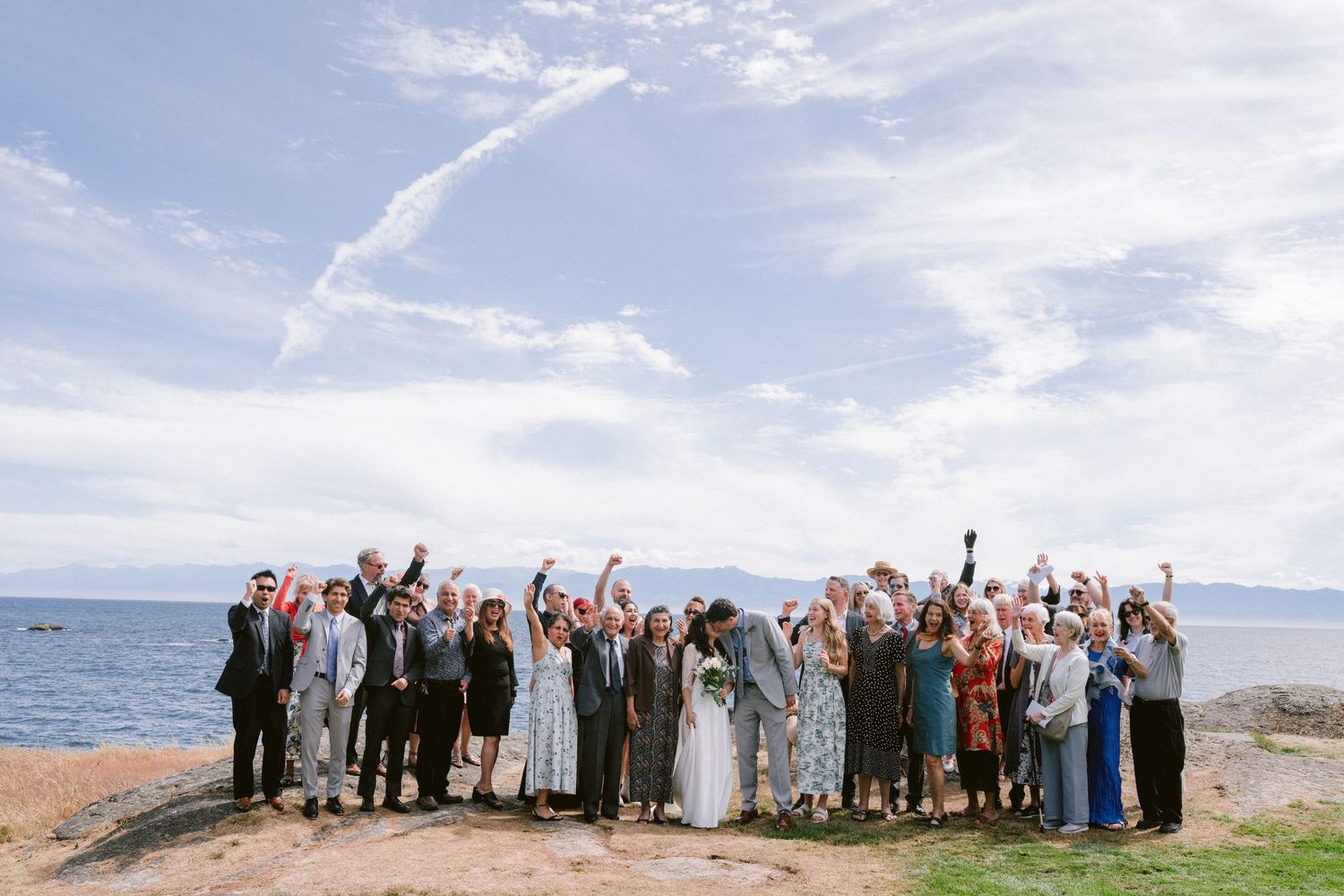 Wedding party celebrating outdoors by the ocean, raising hands in joy on a rocky shoreline under a blue sky.