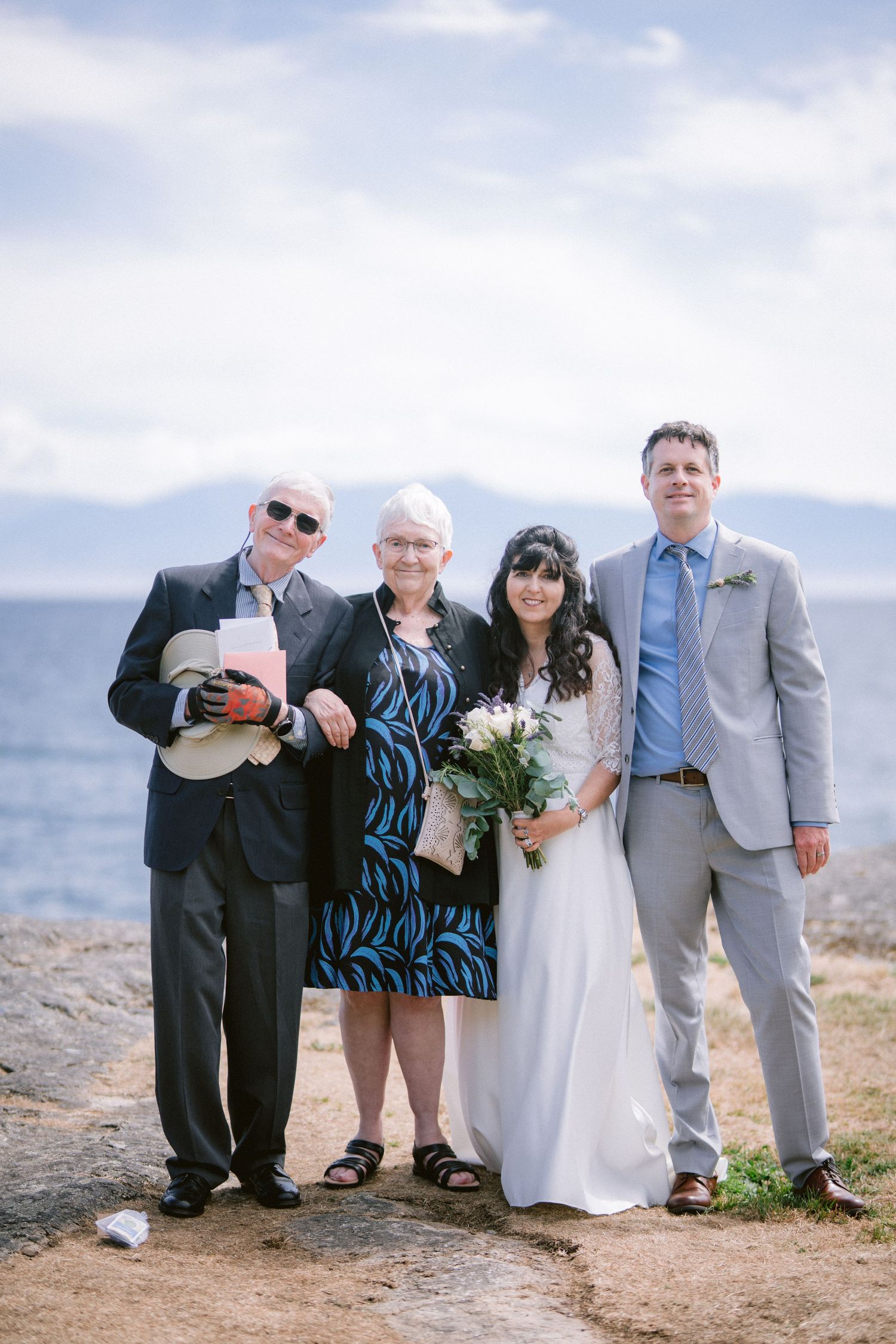 Wedding party of four poses on rocky shoreline with ocean and mountains in background.