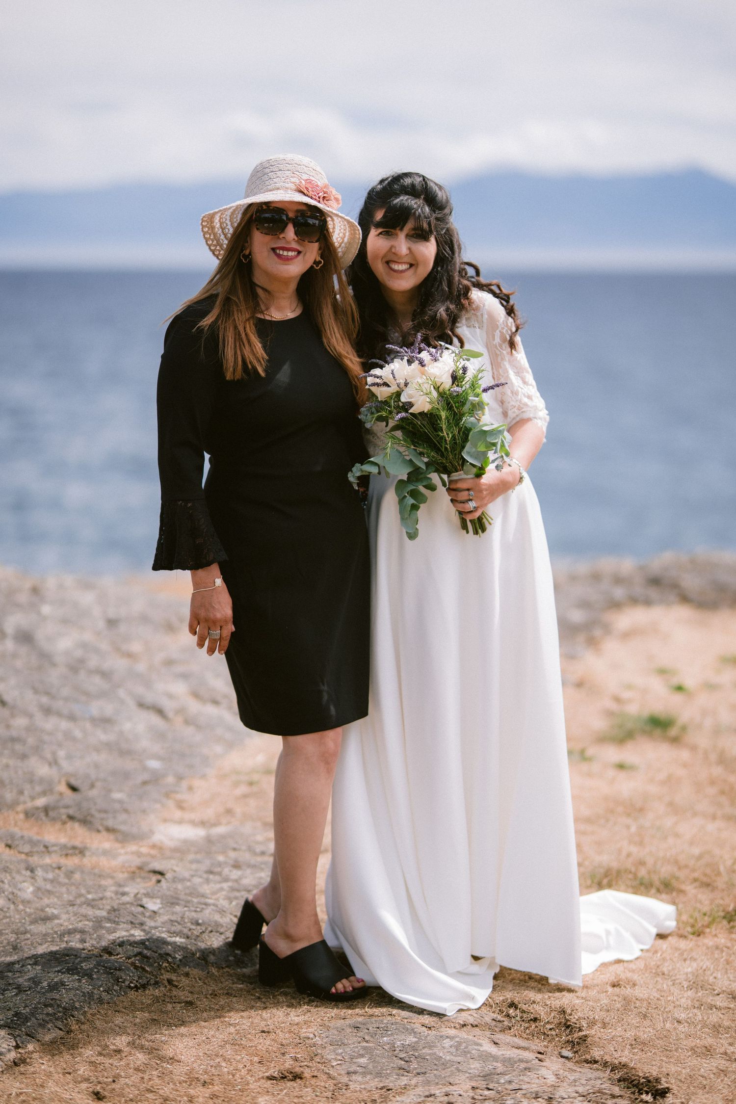 Bride in white gown holding floral bouquet posing with guest in black dress by the seaside.