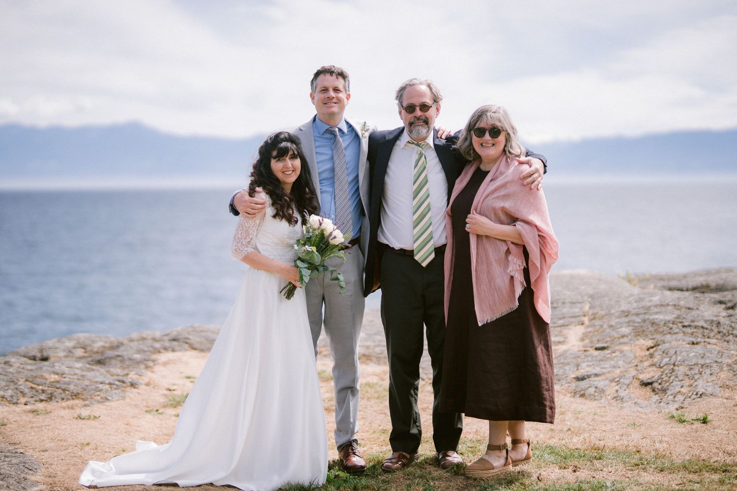 Wedding group portrait of bride, groom, and two guests posing on rocky shore with blue ocean behind them.