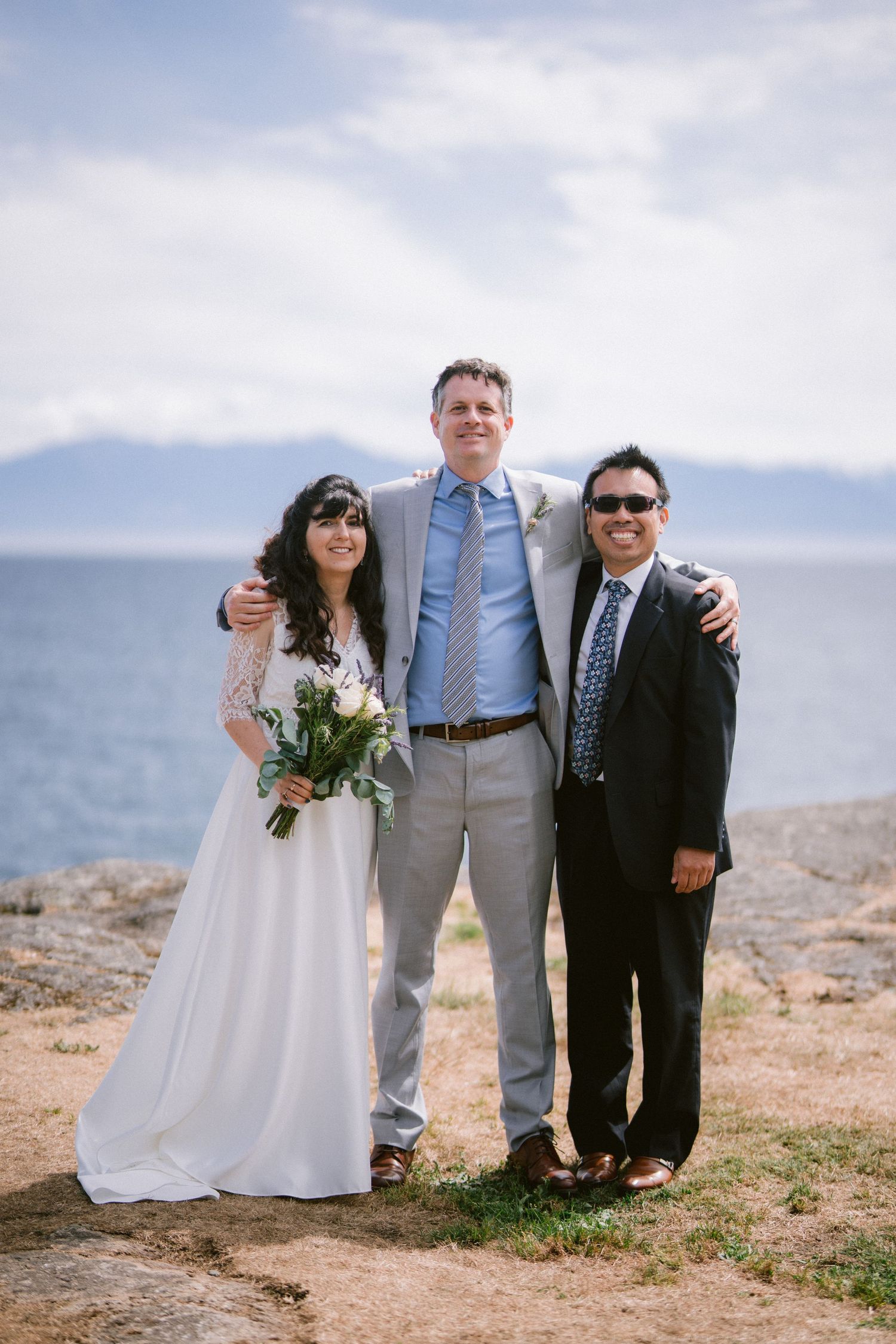 Bride and groom in gray suit posing with tall officiant on rocky coastal bluff with mountain views.