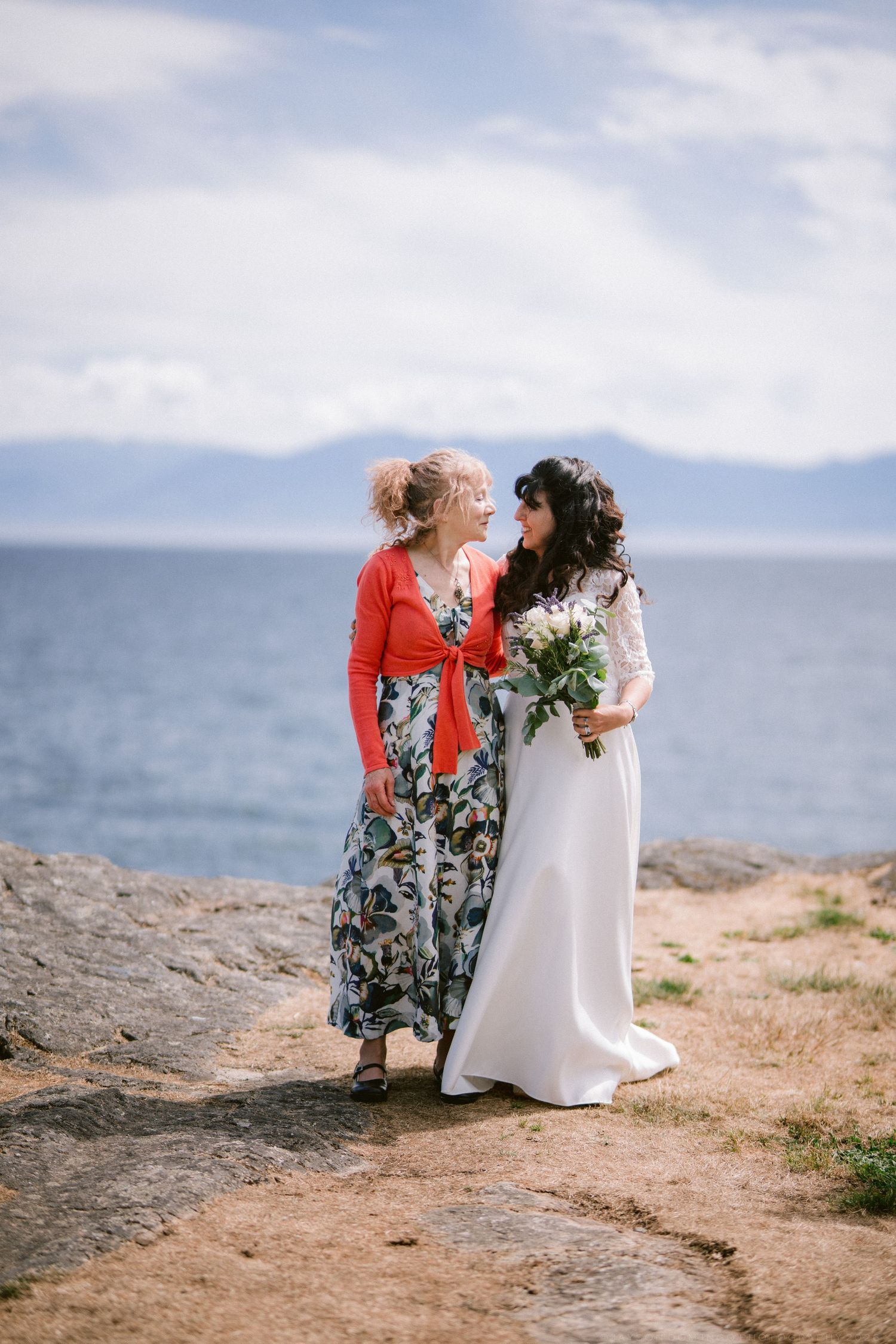 Bride in white lace gown and bridesmaid in floral dress sharing a tender moment by the ocean.