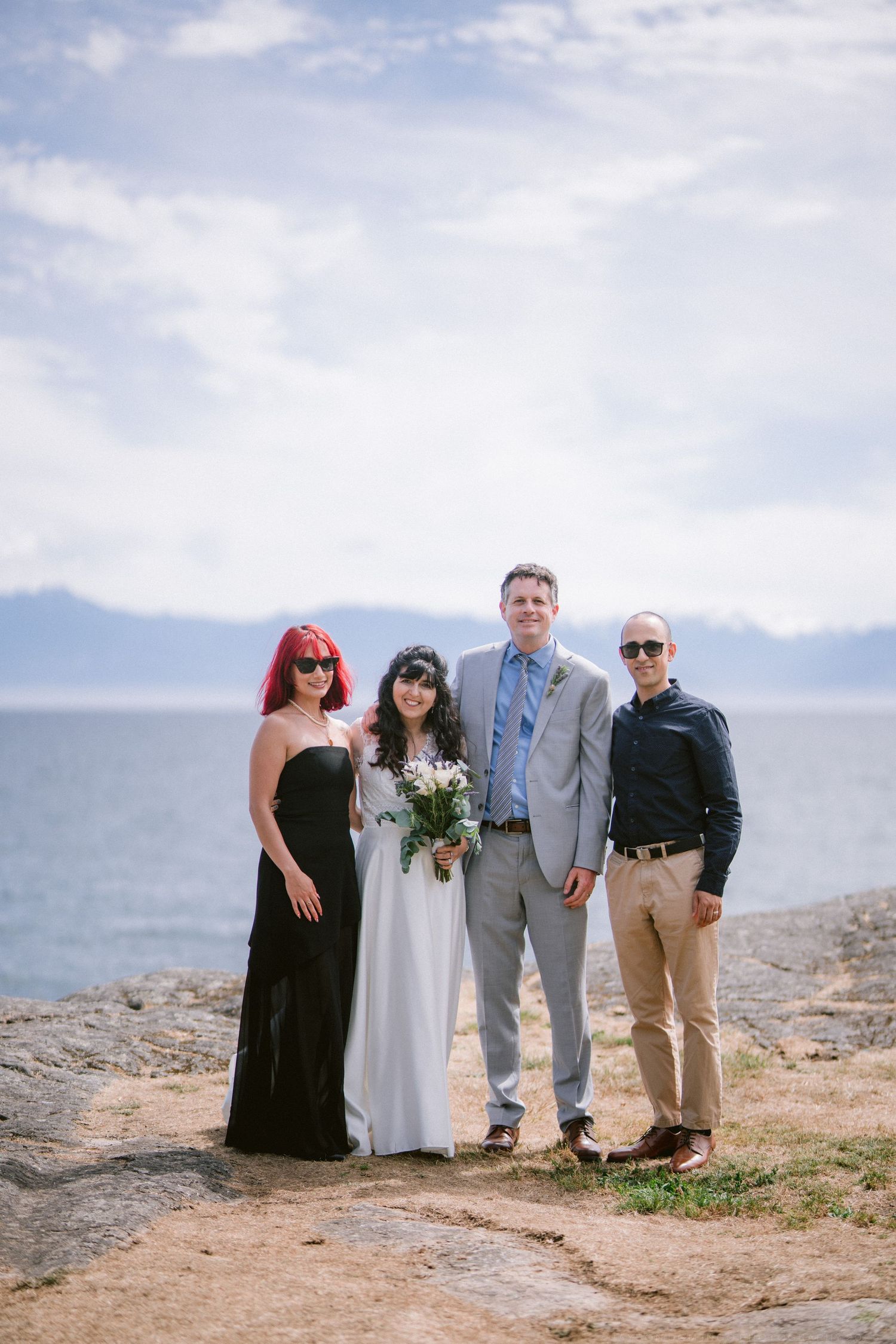 Wedding group of four posing on rocky shoreline with mountains and blue sky in background.