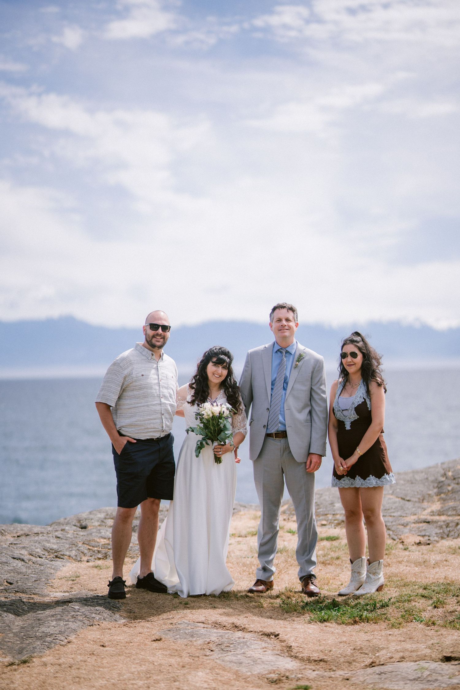 Wedding couple with guests posing on rocky coastal cliff with ocean and mountains behind.