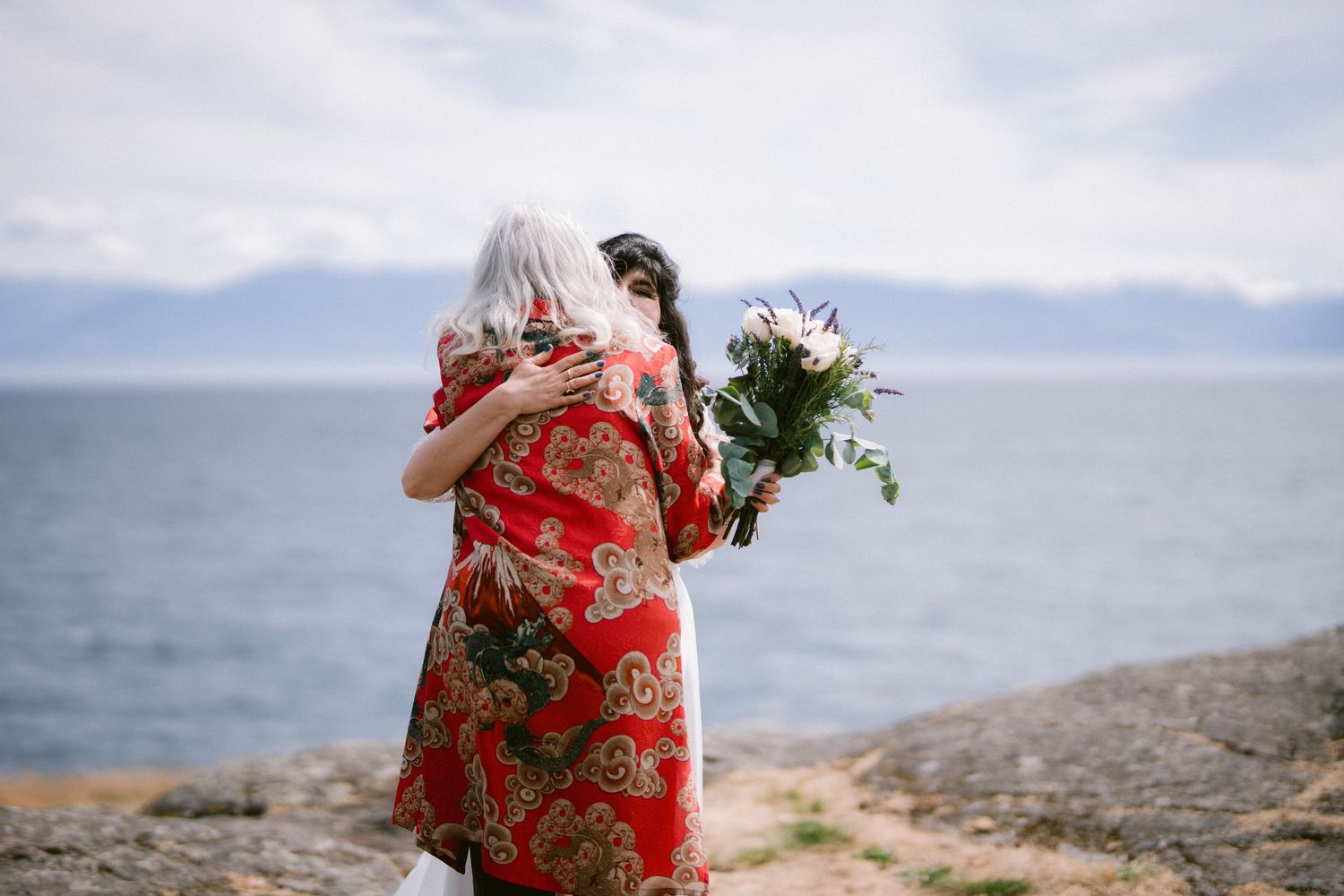 Bride embracing woman in red floral dress on rocky shoreline with bouquet and mountain views in background.