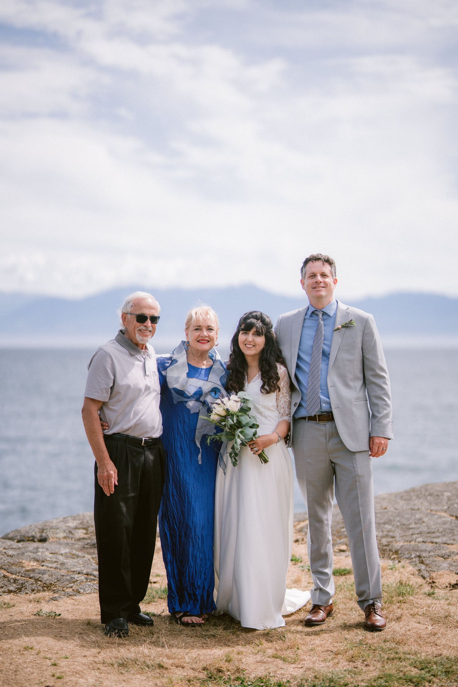 Wedding portrait of bride and groom with two guests near rocky oceanside with mountains behind.