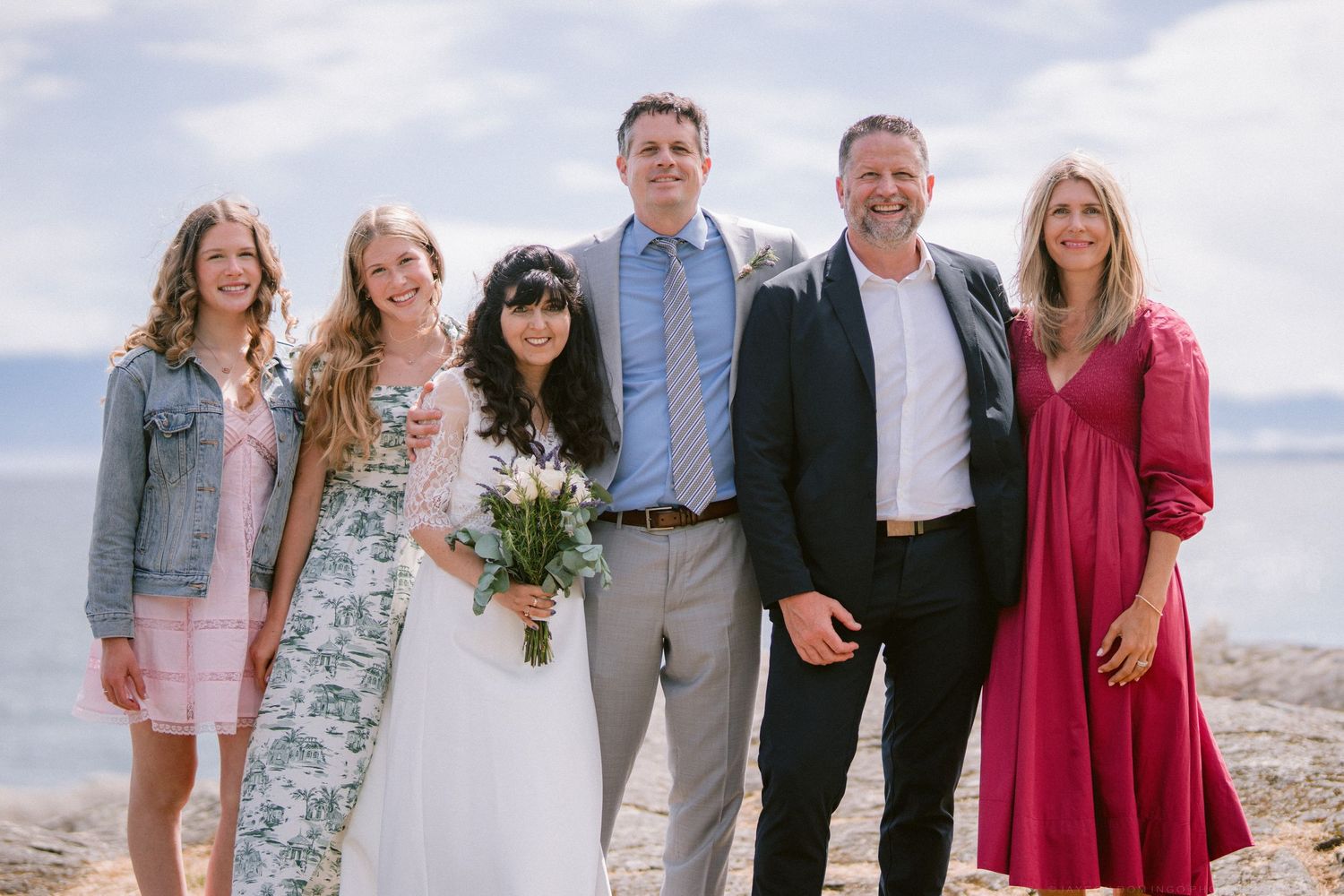 Newlywed couple poses with four family members on a rocky beach shore on their wedding day.