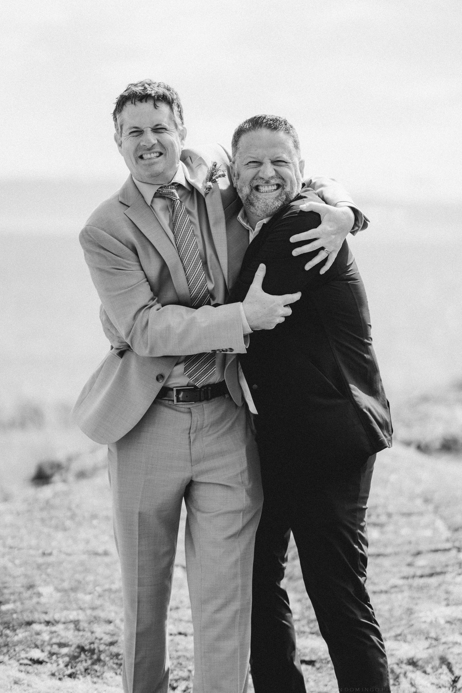 Two men in suits hugging and smiling outdoors in a black and white wedding photo.