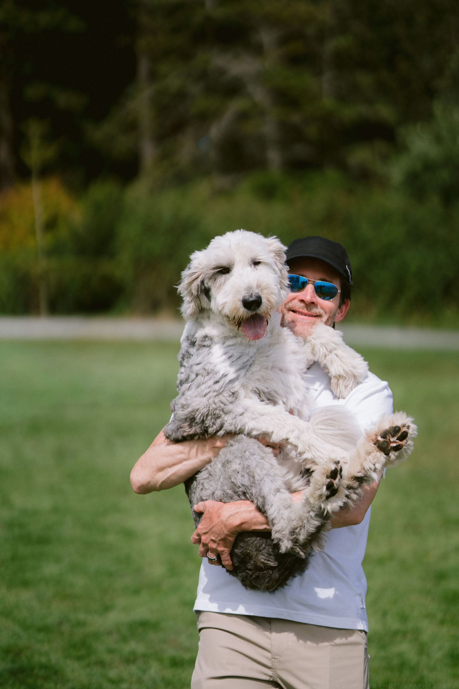 Smiling man in sunglasses and white shirt holding a large fluffy white dog outdoors.