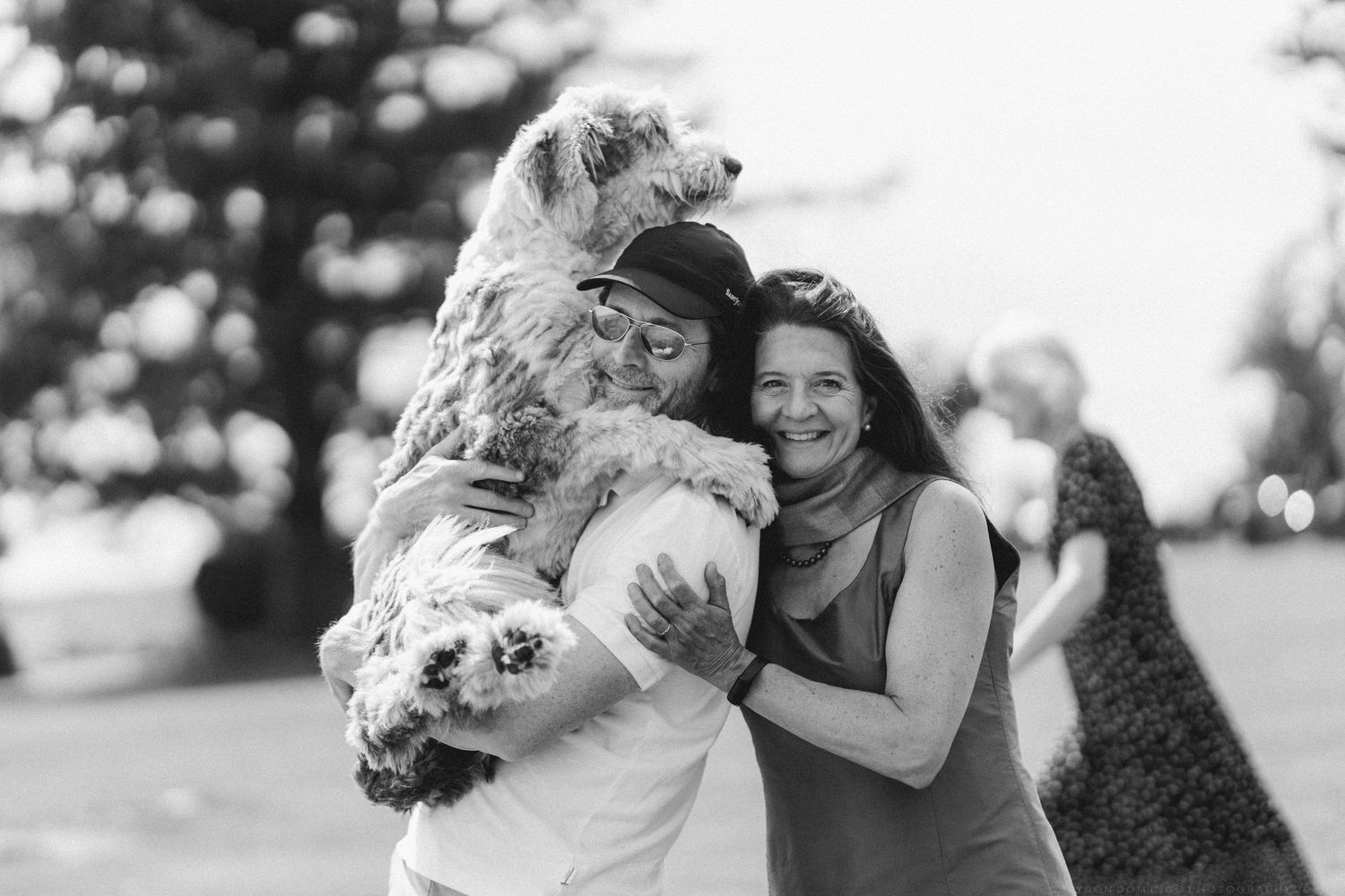 Two women and a fluffy dog share a joyful hug outdoors in a black and white photo.