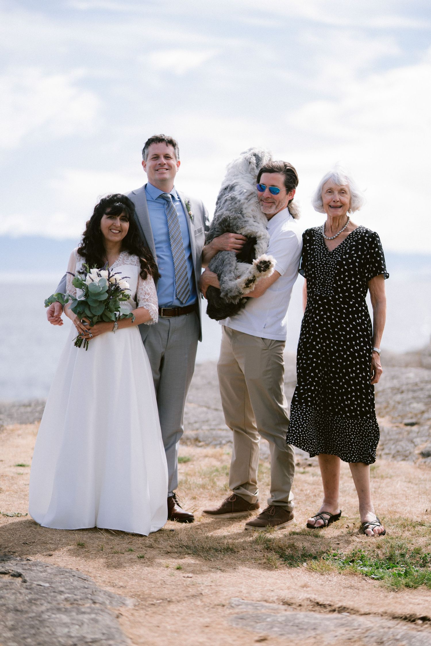 Wedding party of four posing outdoors by the sea, including bride, groom, and guests with a dog.