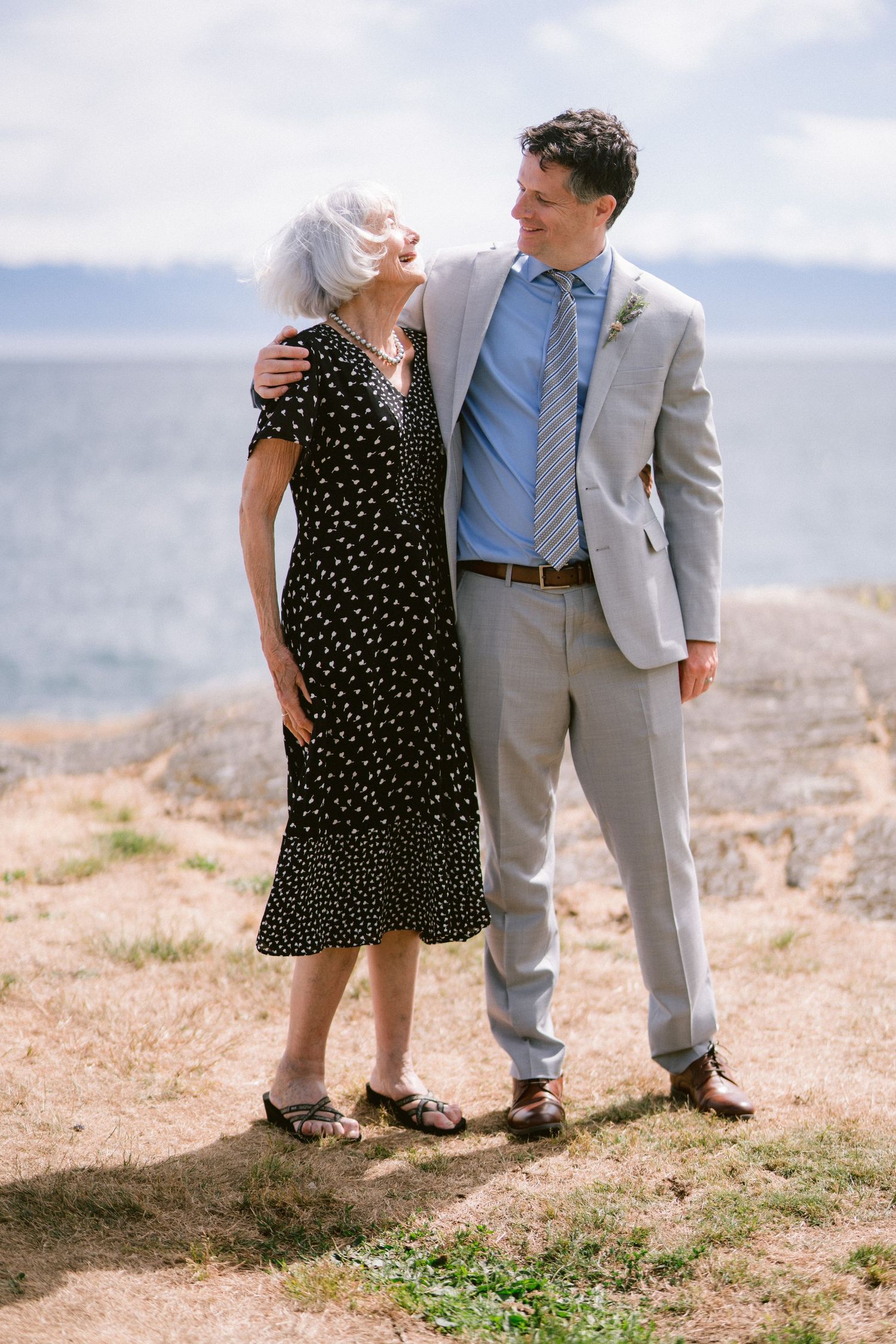 Groom in gray suit embracing an older woman in black dress by the ocean.