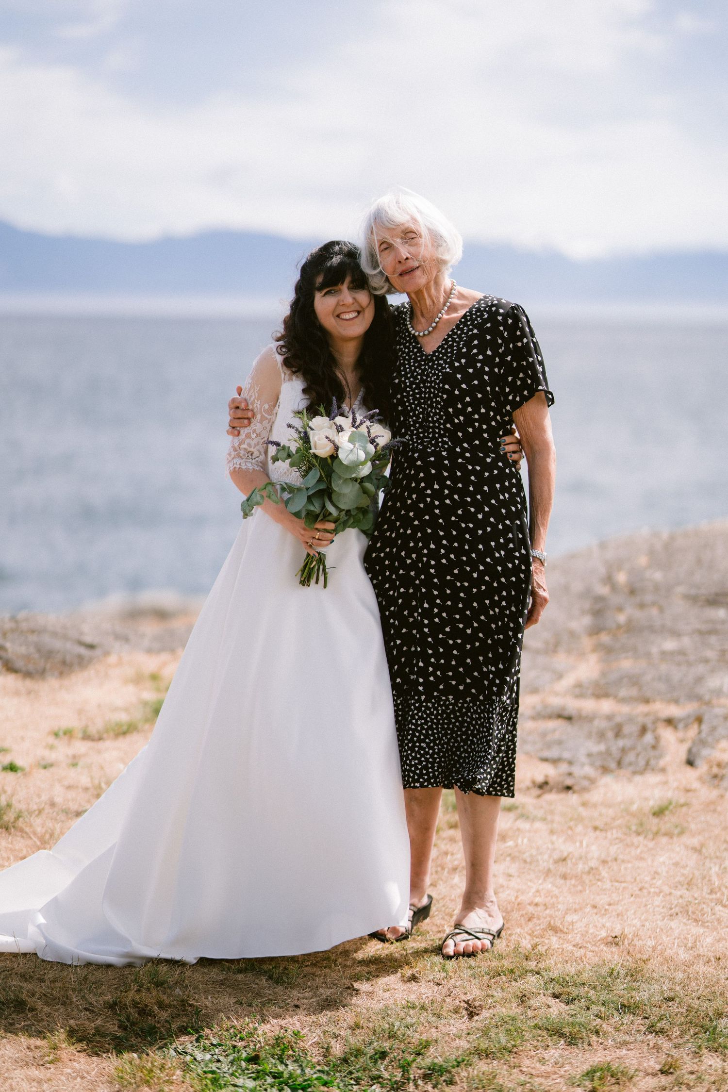 Bride in white lace gown holding bouquet posing with elderly woman in black polka dot dress by the sea.