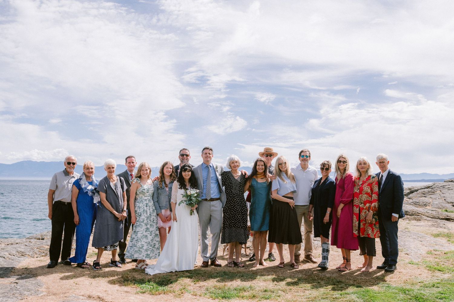 Large wedding group portrait on rocky coastal shoreline with mountains and cloudy sky behind them.
