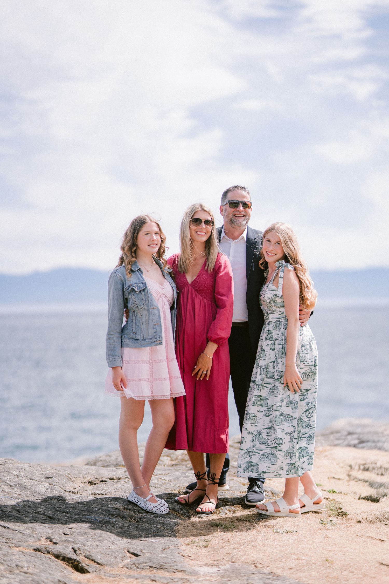 Family of four posing together on rocky shoreline with ocean and cloudy sky backdrop.