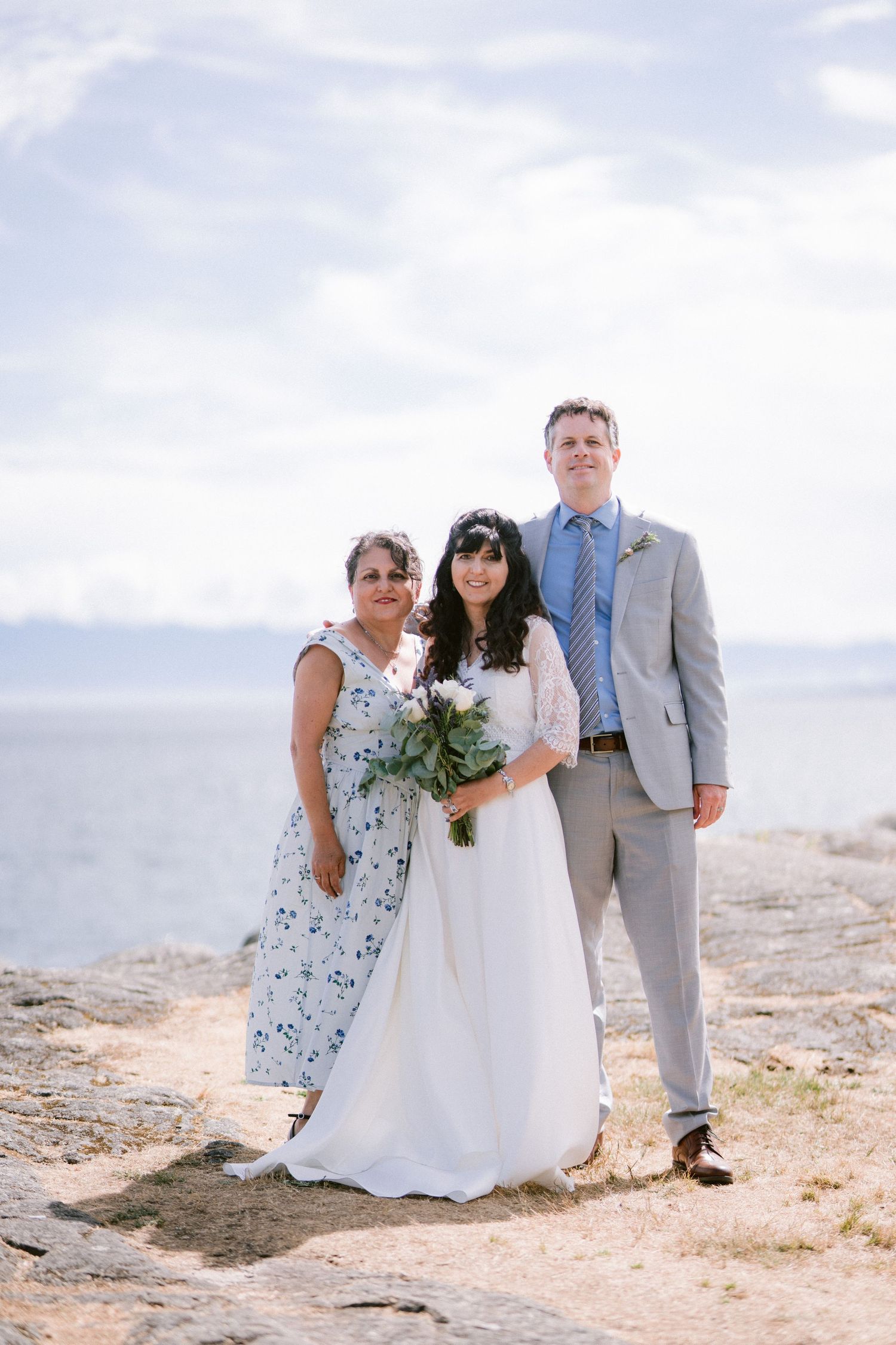 Bride holding bouquet stands with groom in gray suit and woman in floral dress by the ocean.