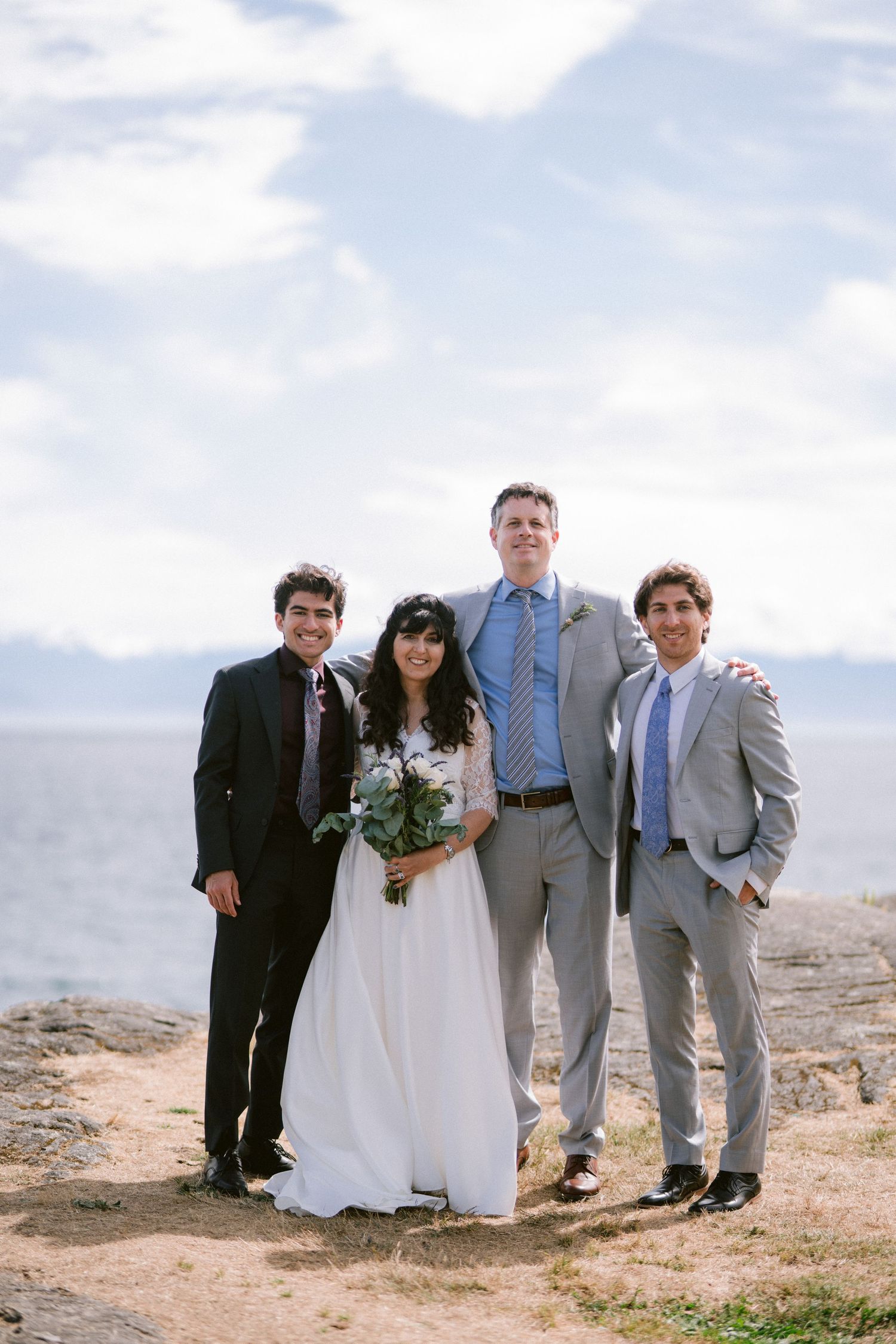 Wedding party of four posing on rocky coastal overlook with ocean and mountains in background.
