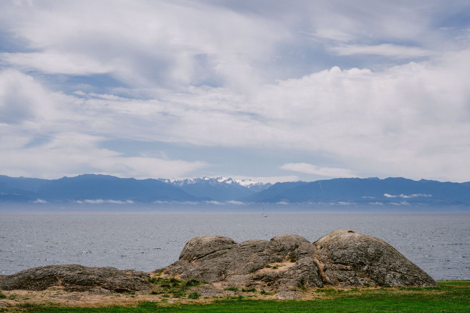 Rocky shoreline with calm ocean waters and snow-capped mountains visible under a cloudy sky.