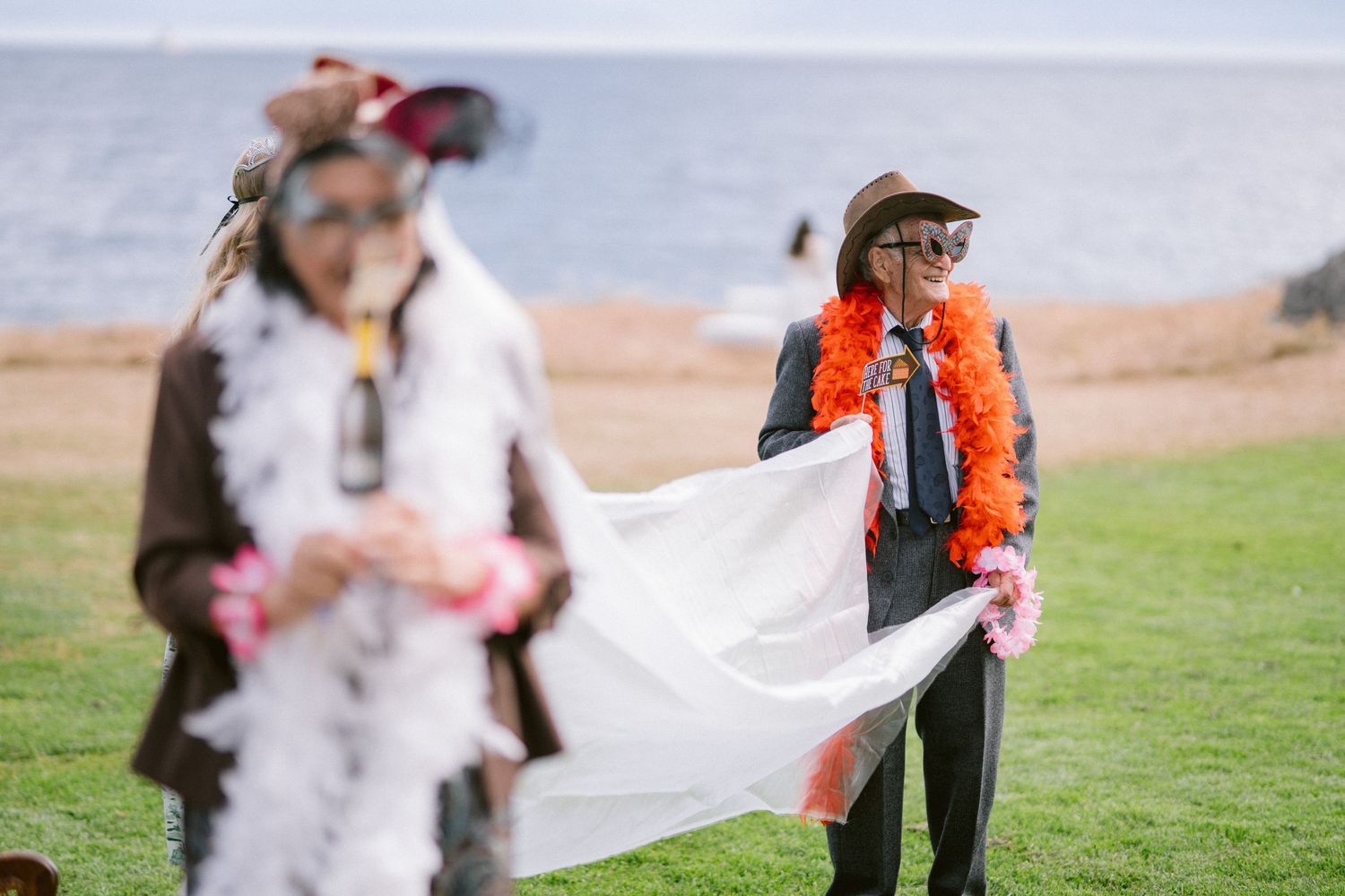 Wedding guests in festive costumes playing with a white veil on a seaside lawn.