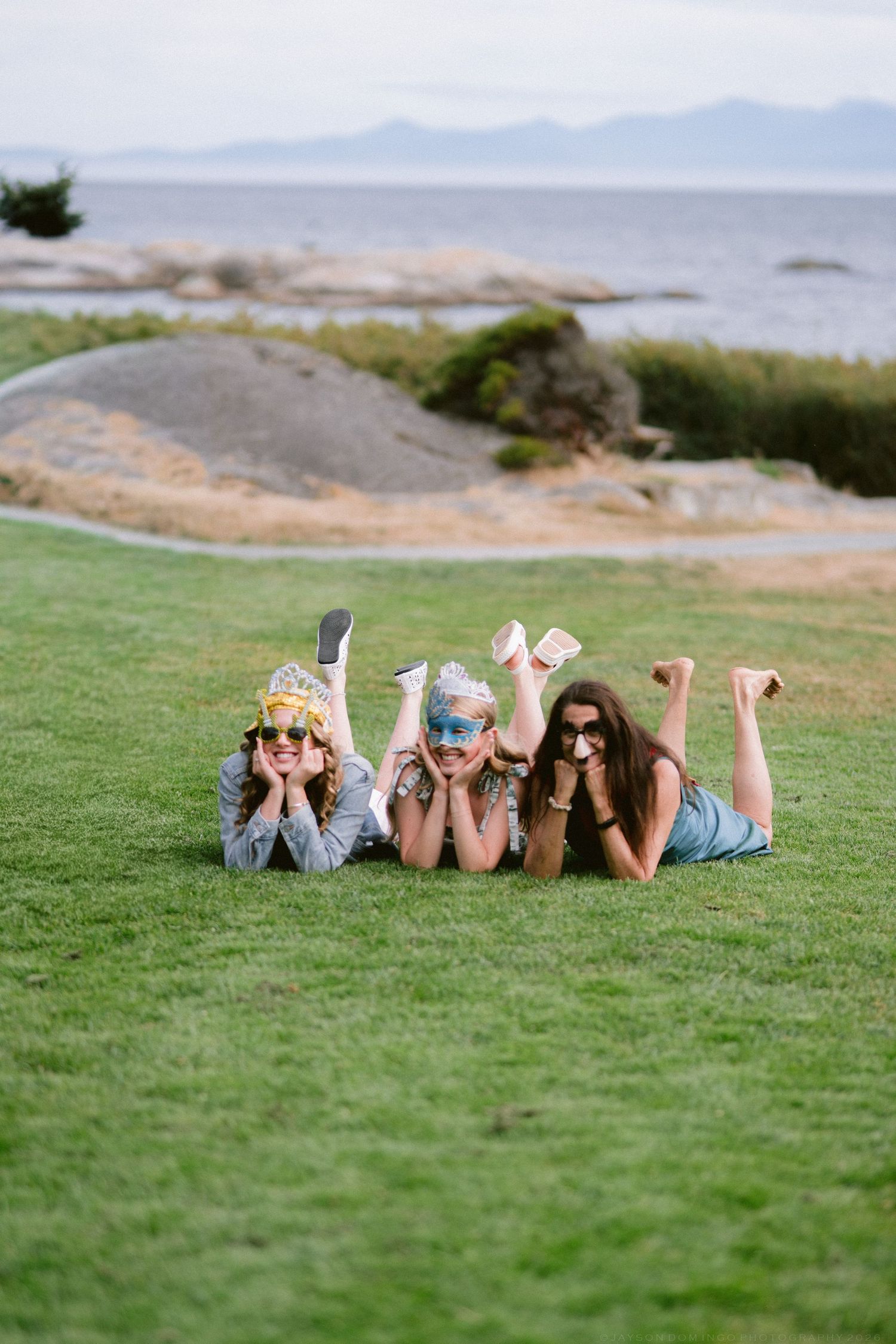 Three girls lying on green grass near the ocean, wearing sunglasses and smiling.
