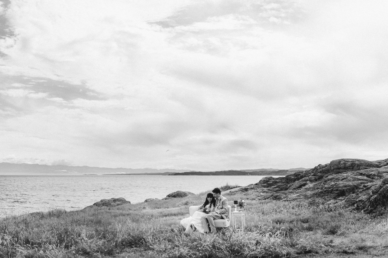 Black and white photo of a couple sitting together on a grassy coastal landscape with ocean views.