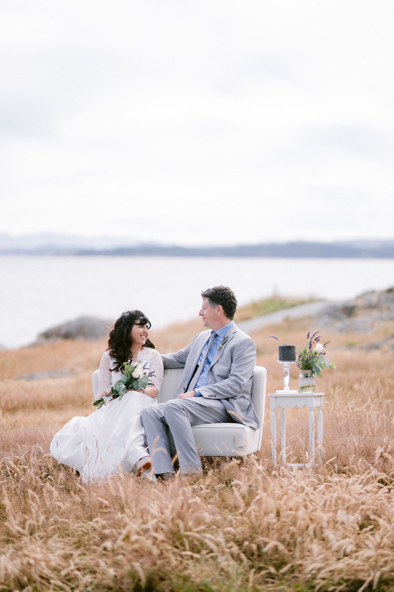 Bride and groom sit together in chairs in golden field with ocean view on wedding day.