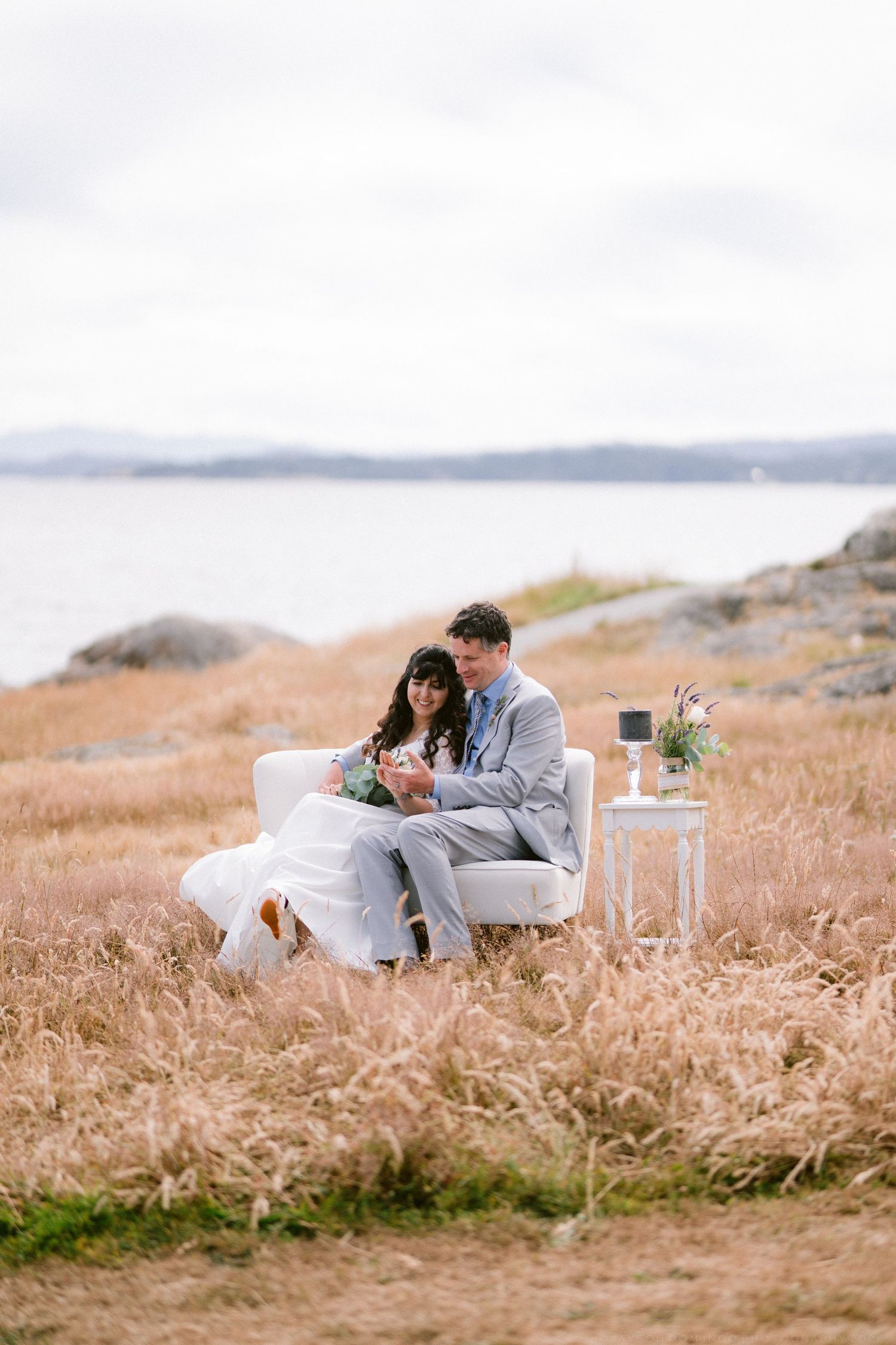 Bride and groom sitting together in a white chair surrounded by golden grass fields near the ocean.
