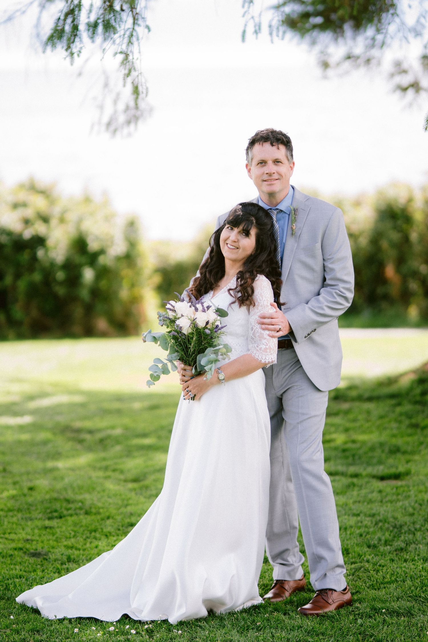 Bride and groom in gray suit embracing on green lawn, bride holding white and pink rose bouquet.