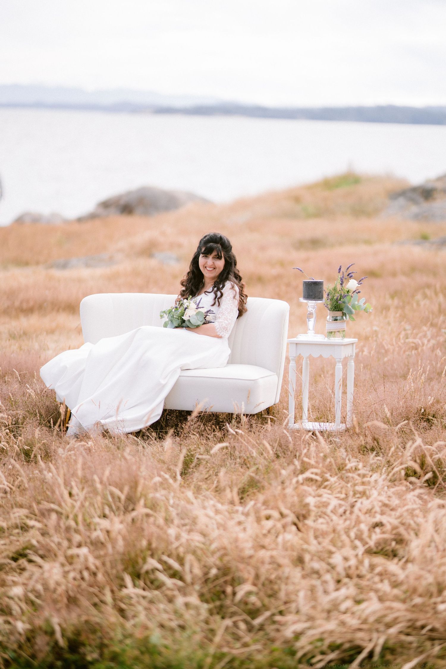 Bride in white gown sitting on white sofa in golden field with bouquet and floral side table by the sea.