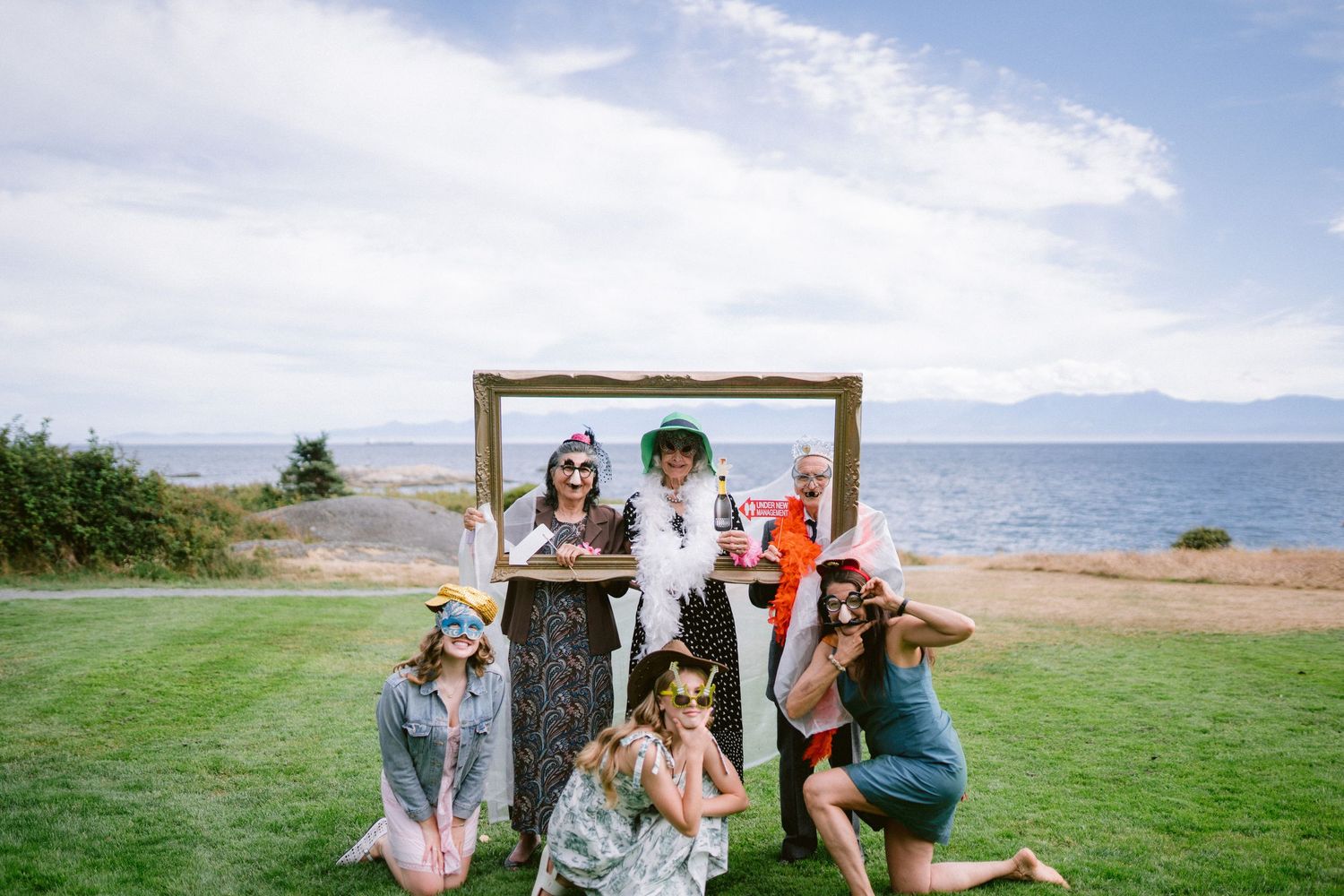 Group of friends posing with a decorative gold frame outdoors near the ocean, wearing fun accessories and costumes.